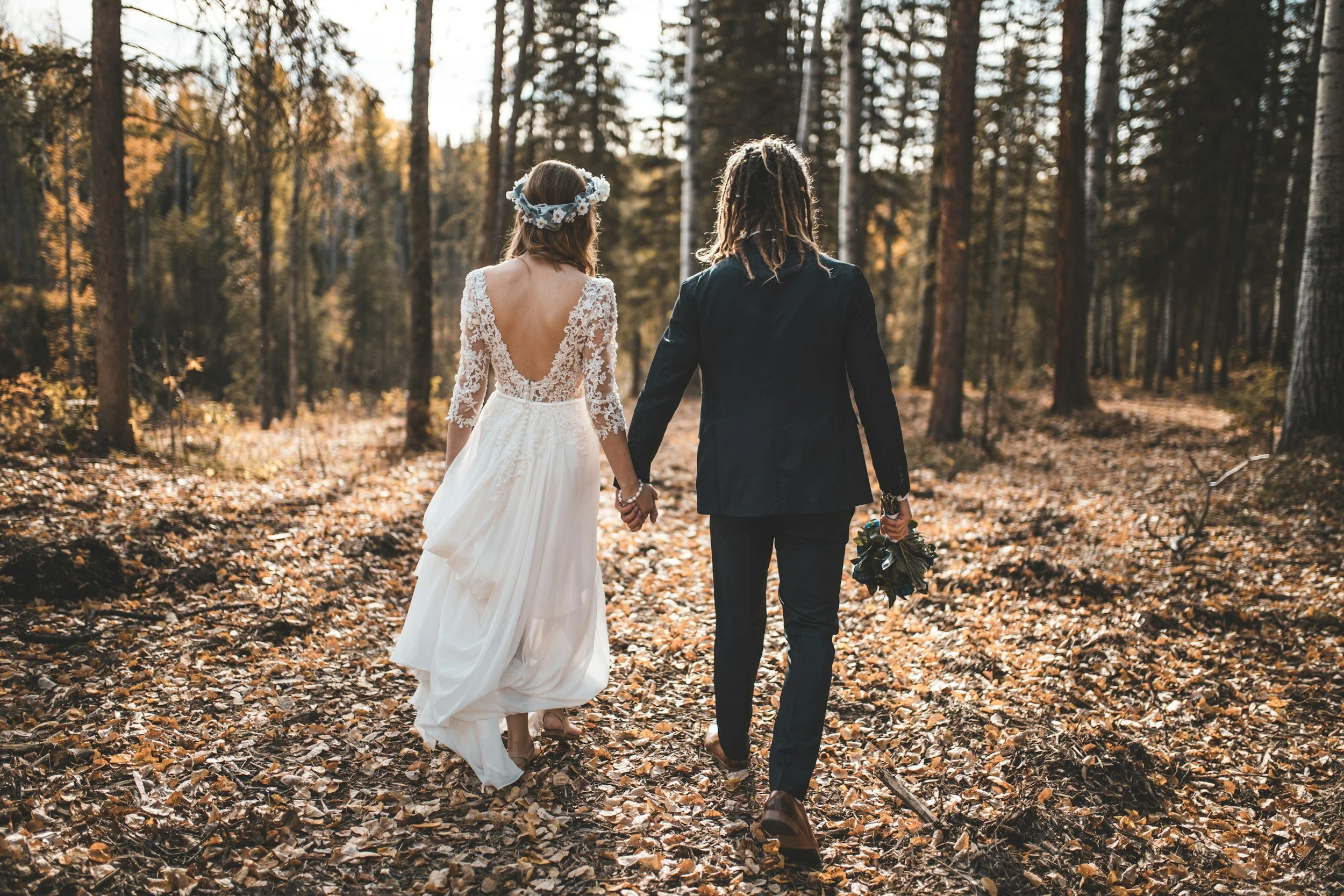 A couple, dressed in wedding attire, walking hand-in-hand through a forested area with fall leaves on the ground.