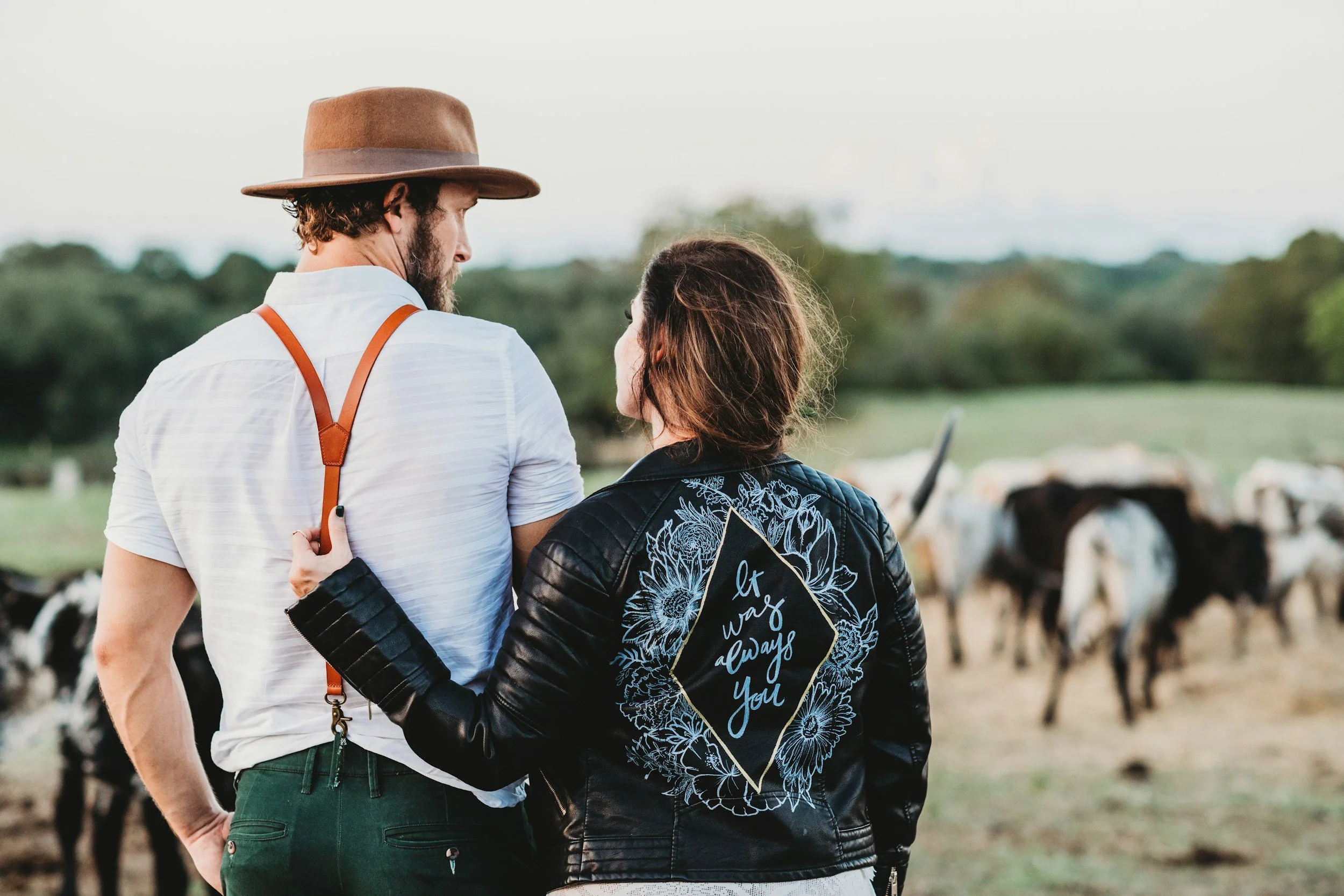 A couple stands close together outdoors in a field with cows in the background. The man wears a brown hat, white shirt, and suspenders. The woman has a leather jacket with floral embroidery and a message that reads, 'It was always you.'