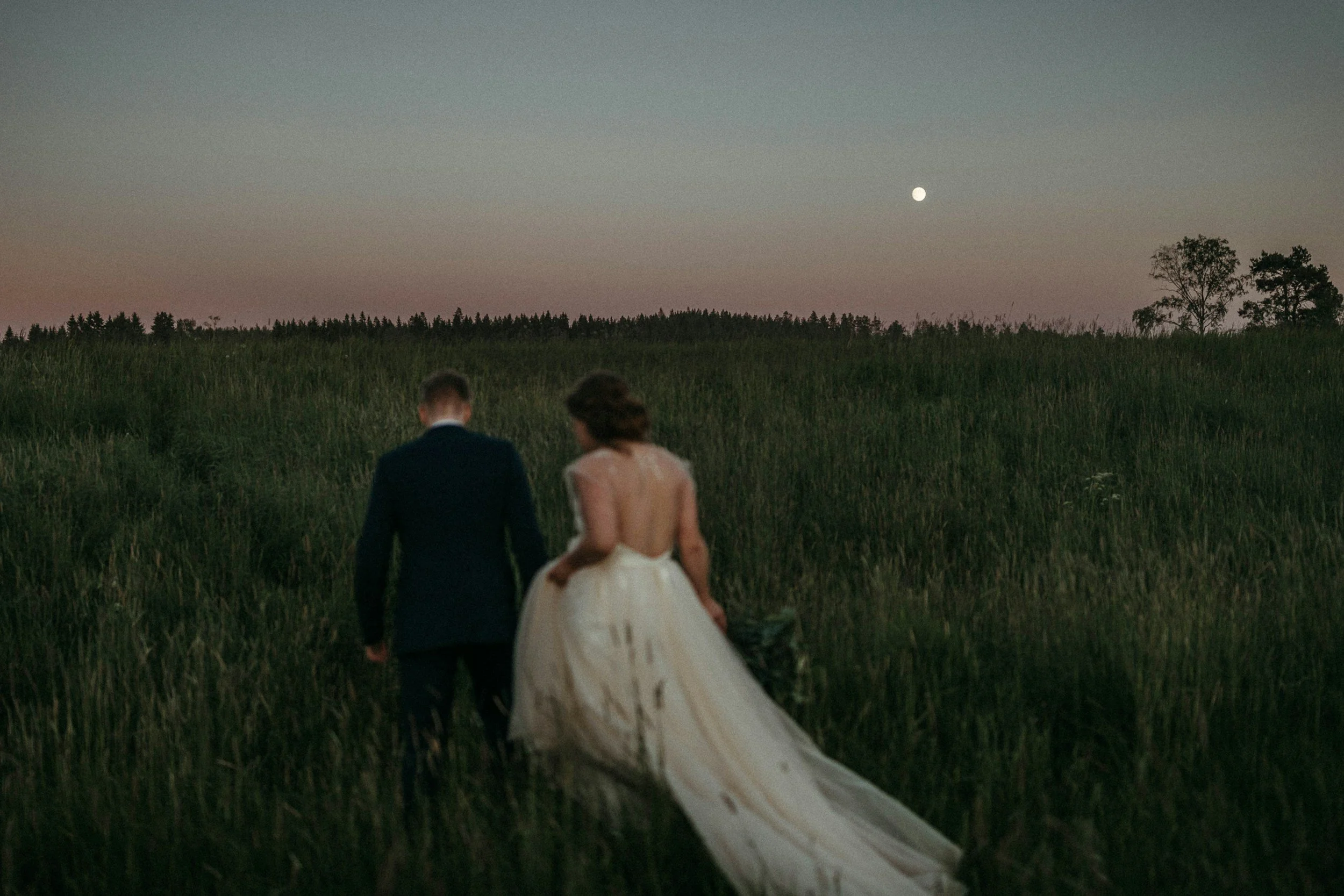 A bride and groom walking through a grassy field during sunset or dusk with a visible moon in the sky.