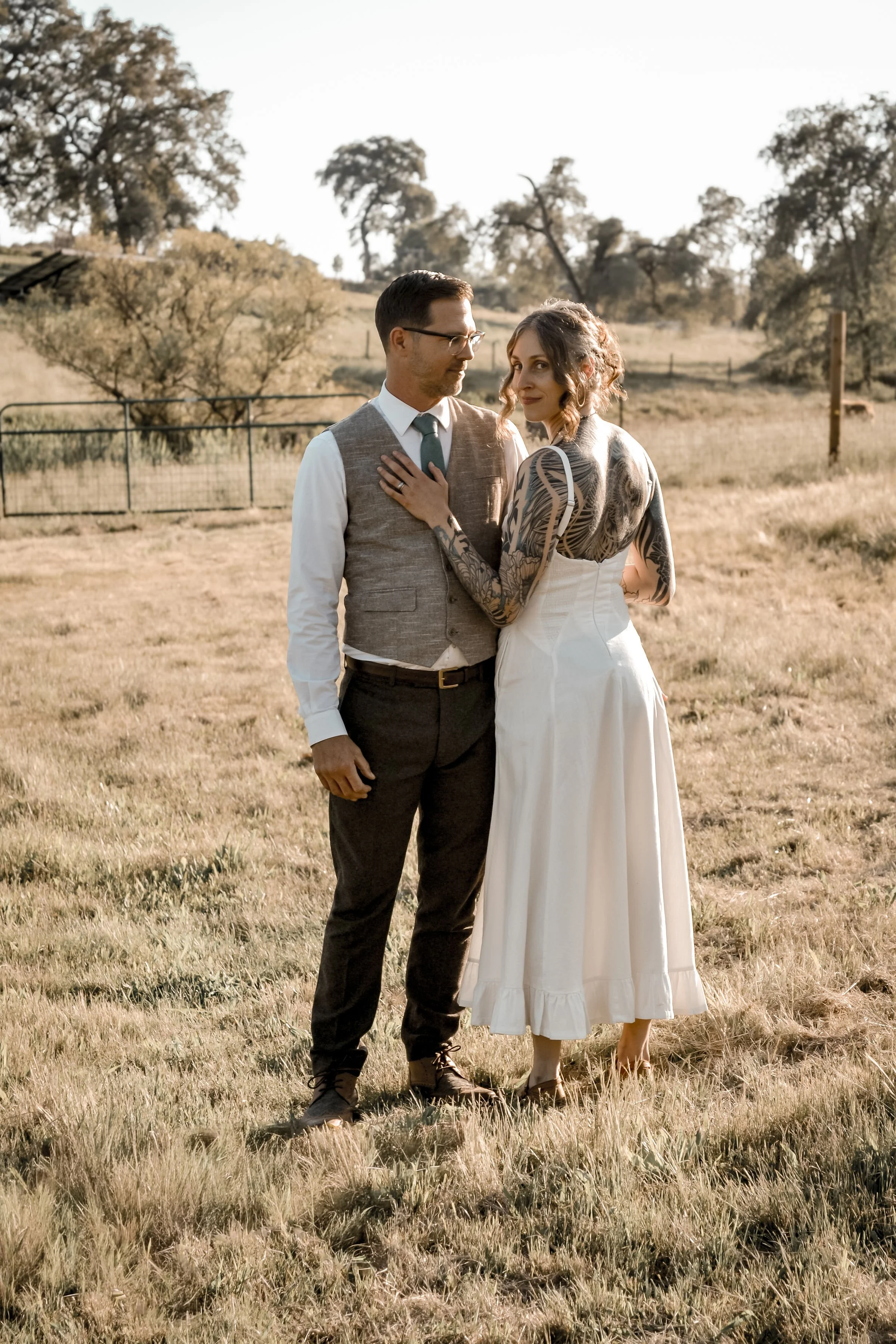 A couple standing in a grassy field with trees in the background during daylight, the woman wears a vintage-inspired white dress and has tattoos, while the man is dressed in a vest, shirt, tie, and dark trousers.