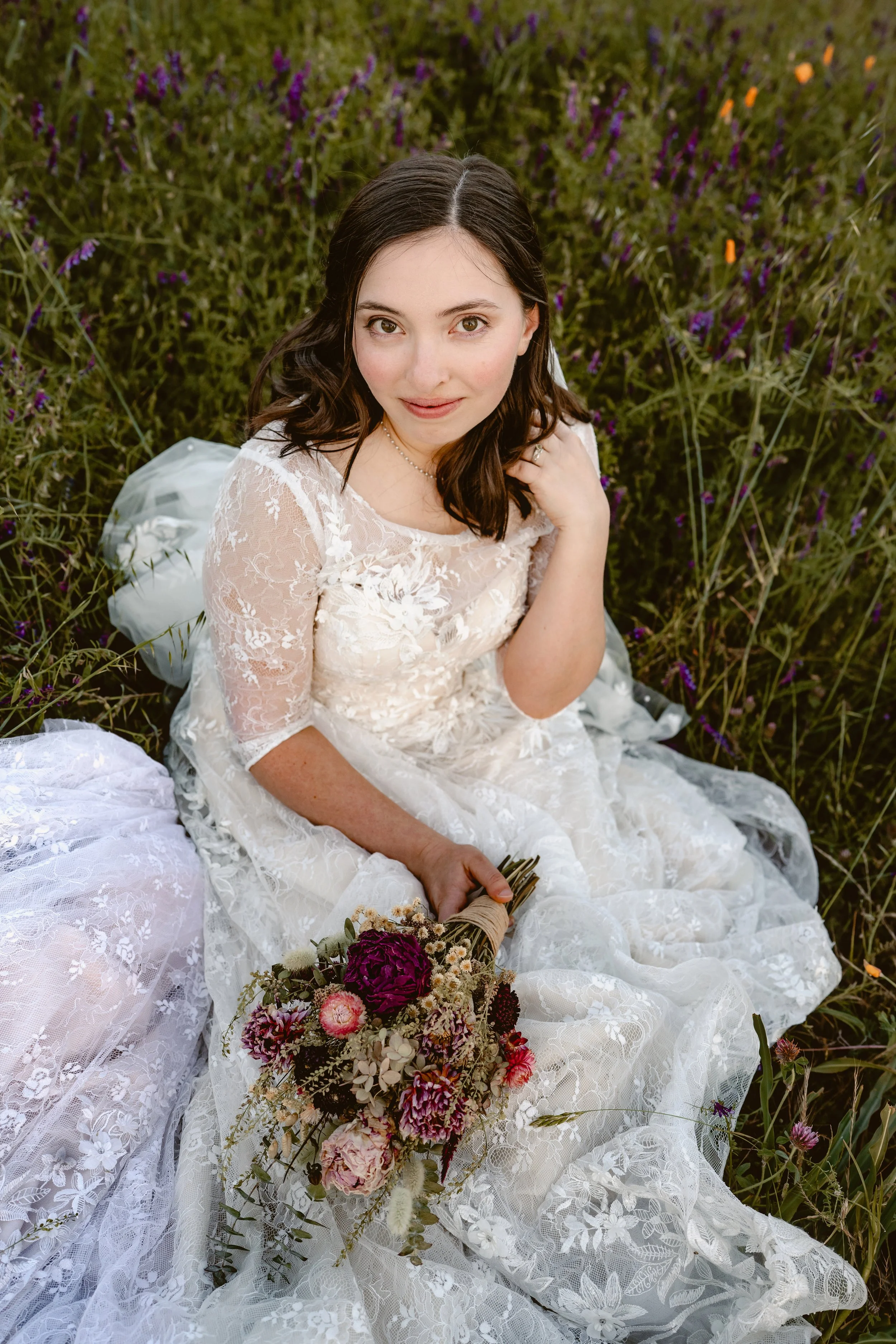 A woman in a white lace wedding dress sitting in a field of purple flowers, holding a bouquet of mixed flowers.