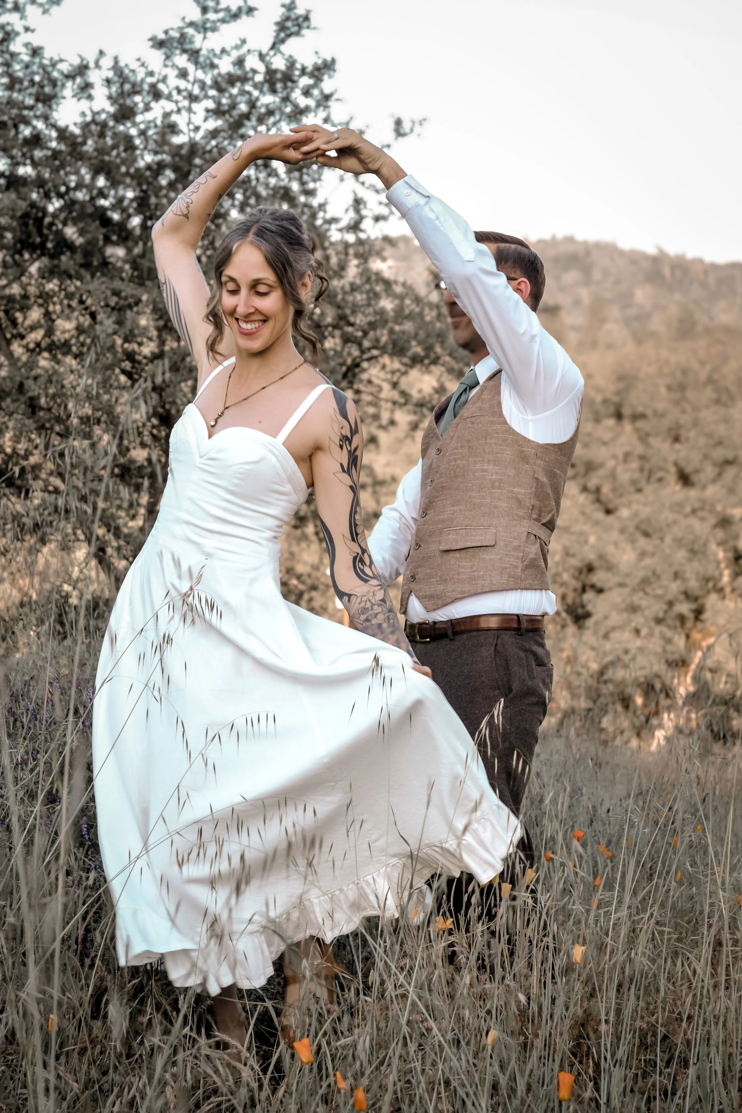 A couple dancing outdoors in a field of tall grass and wildflowers, with a background of trees and distant hills, during sunset.