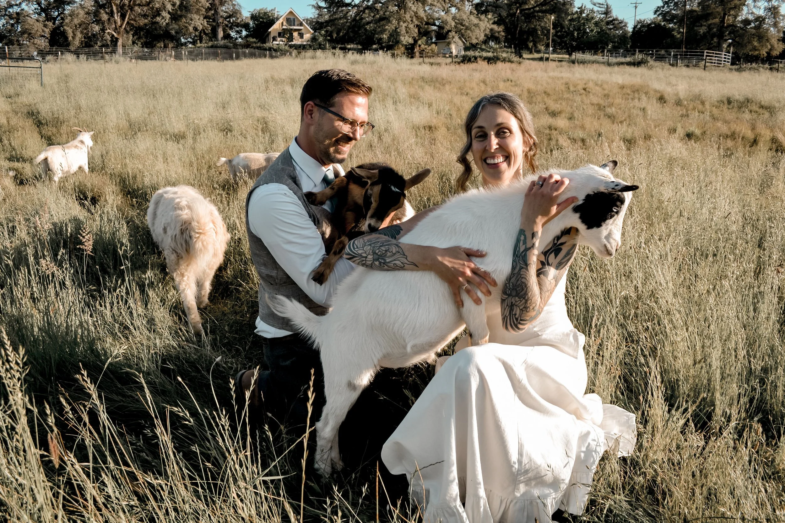 A man and woman outdoors in a grassy field, smiling and holding a white and black goat. Several other goats are grazing in the background.