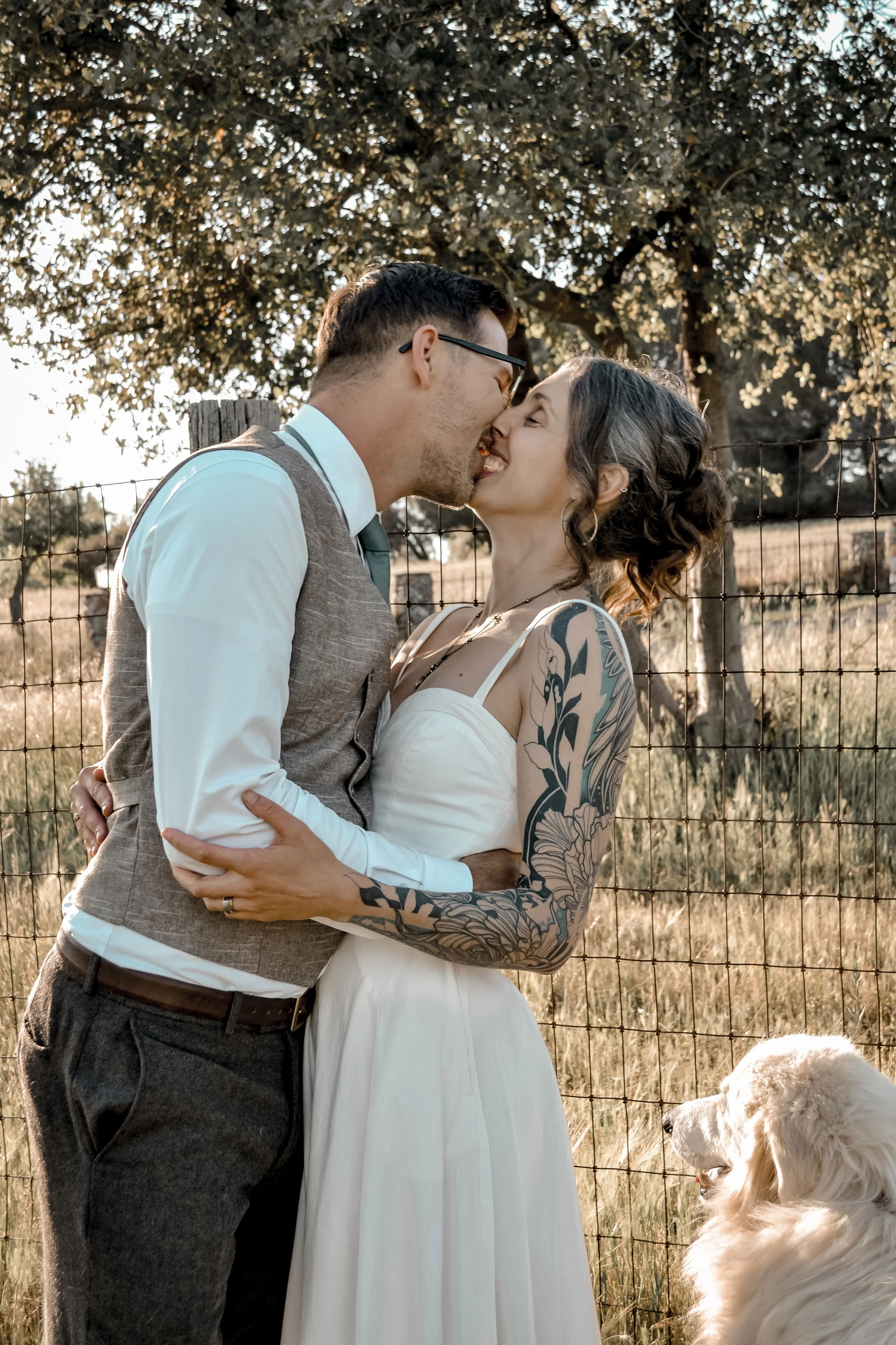 A couple kissing outdoors near a wire fence, with trees and a field in the background. The woman has tattoos on her arm and shoulder, and the man is wearing glasses and a vest.