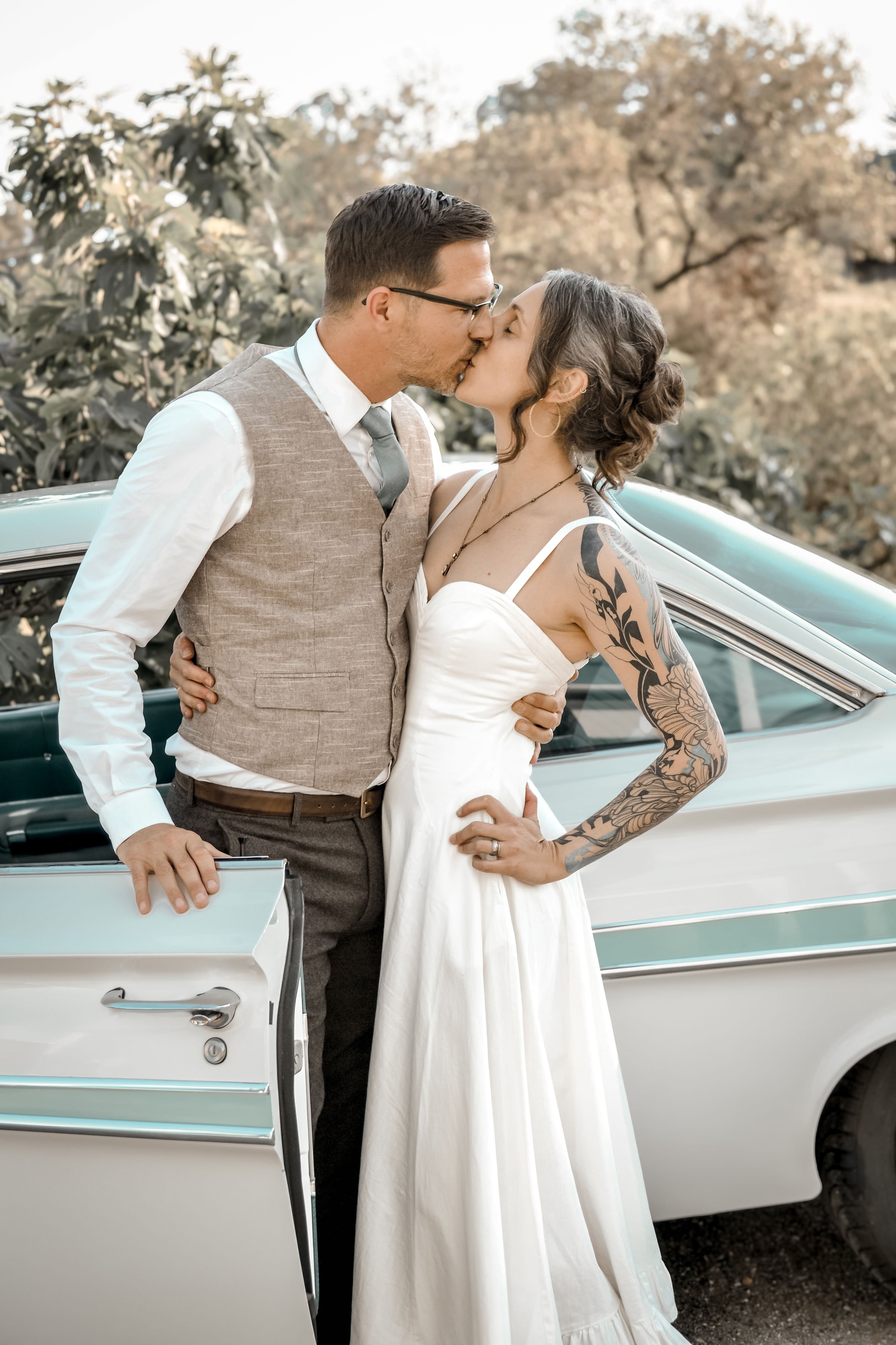 A couple, dressed in wedding attire, sharing a kiss in front of a vintage white car outdoors with autumn trees in the background.