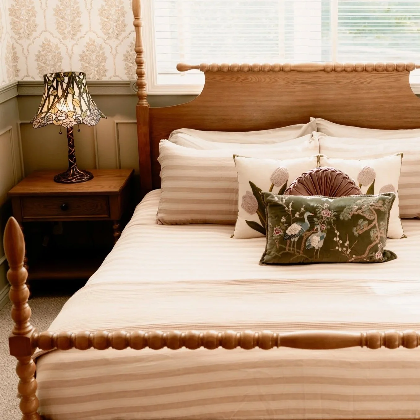 A bedroom with a wooden bed frame, striped cream bedding, three decorative pillows including one embroidered with birds, a wooden nightstand, and a Tiffany-style table lamp, with a window with blinds in the background.