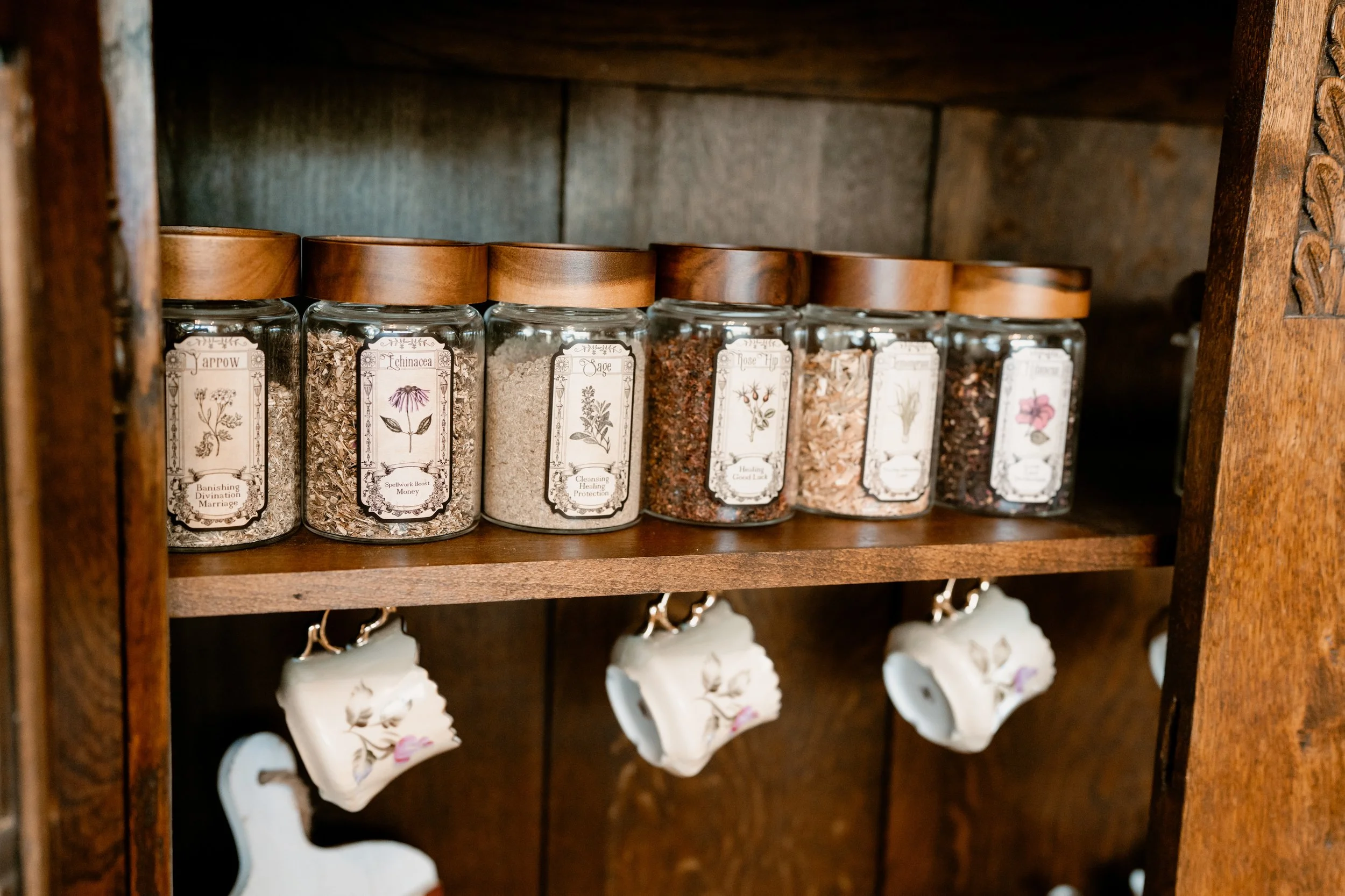 A wooden shelf holding glass jars with wooden lids, labeled with different herbs and spices, and small ceramic cups hanging below, part of a kitchen or herb storage area.