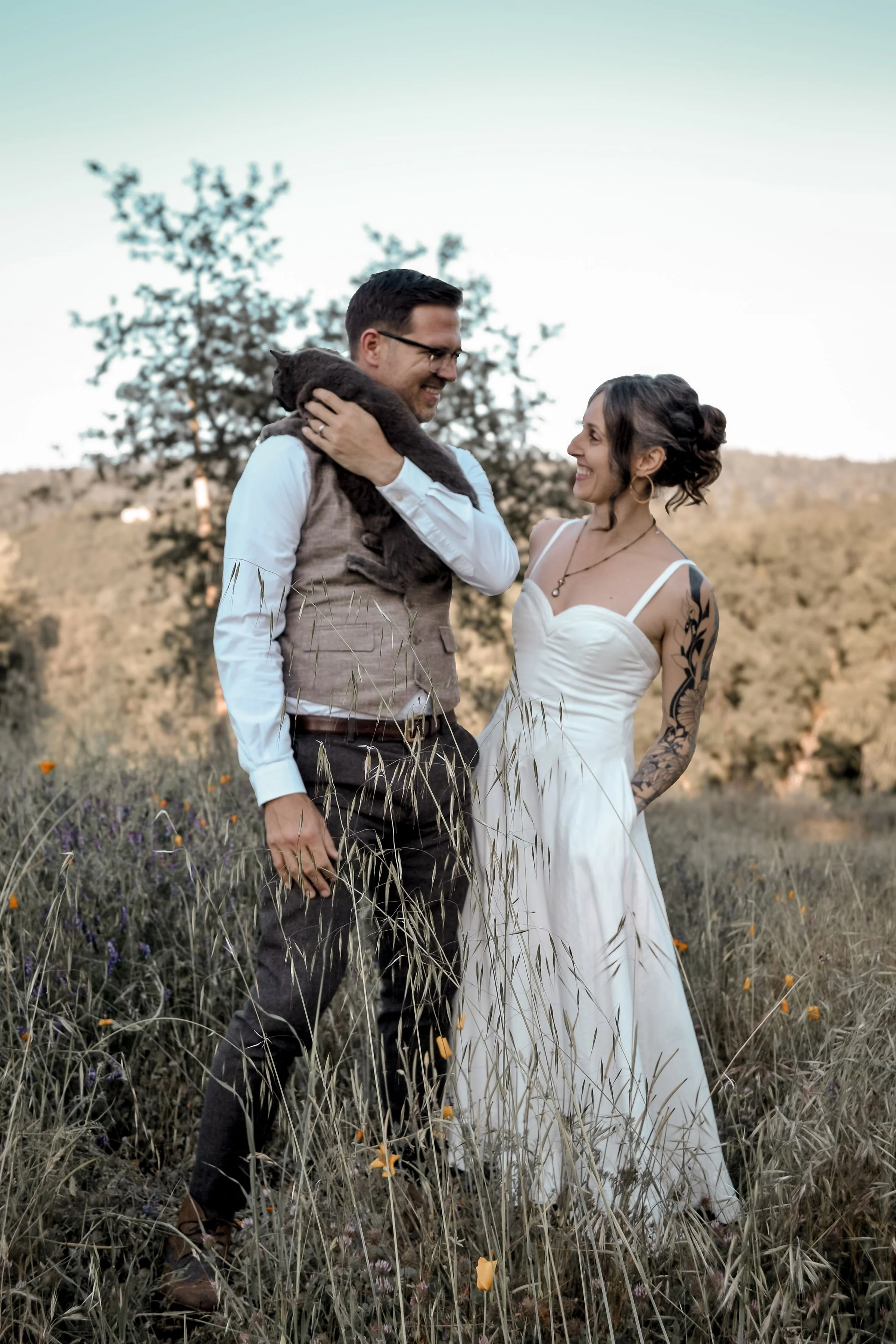 A smiling couple in wedding attire standing in a field of tall grass and wildflowers, with a hillside and trees in the background, during what appears to be late afternoon or early evening.