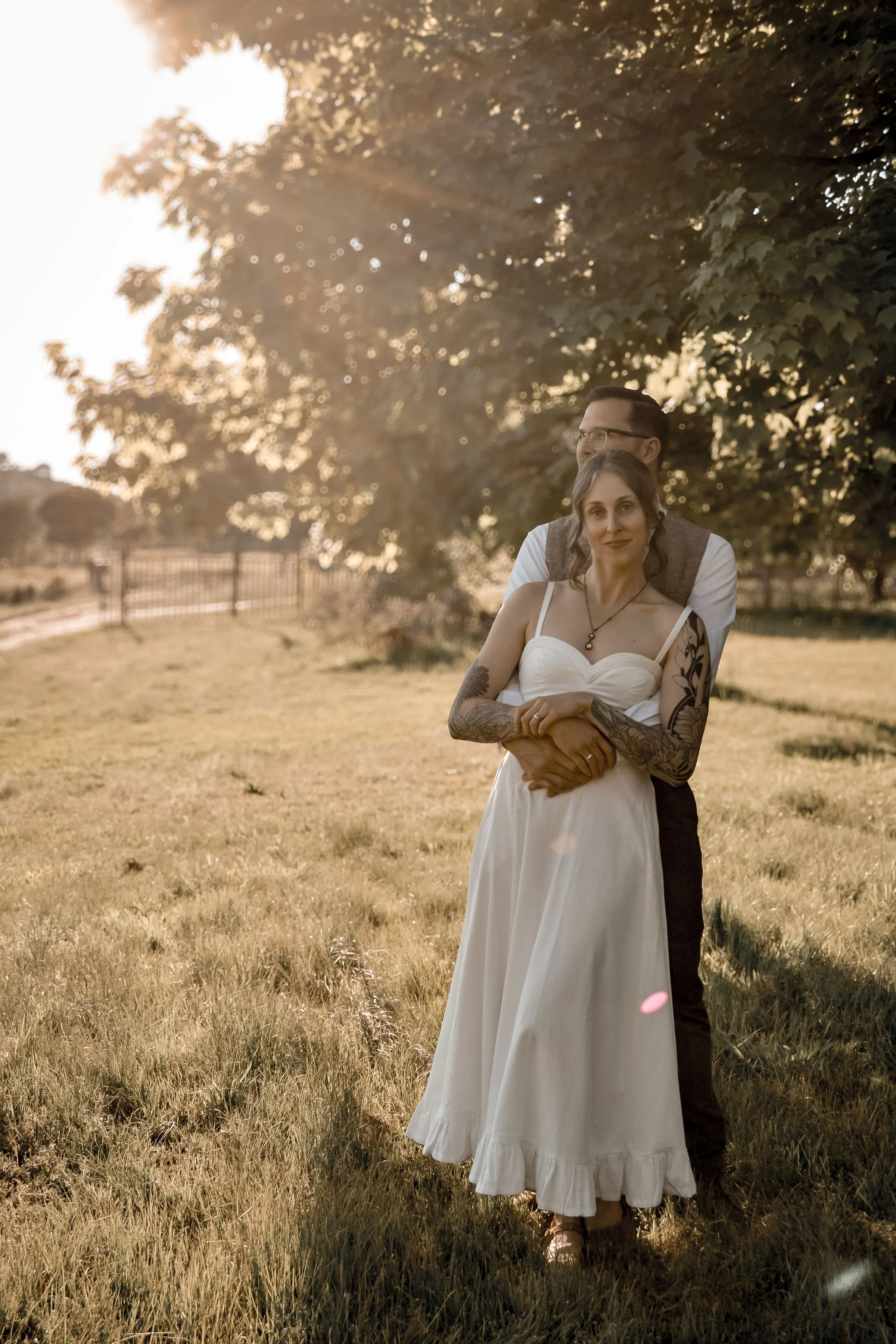 A couple stands outdoors in a grassy field with trees and a fence in the background, during sunset. The woman in the foreground has tattoos on her arms and is wearing a white dress. The man behind her is wearing glasses, a vest, and a white shirt, an