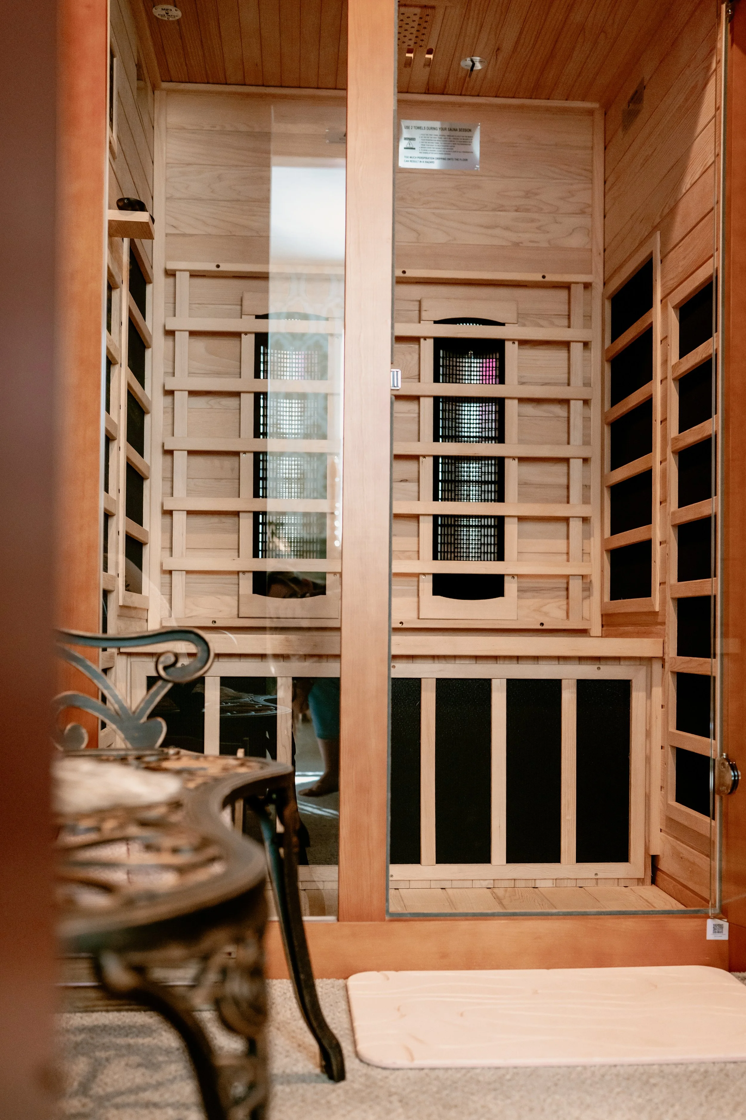 A wooden sauna interior with a glass door, a black vent, and wooden slats. There is a white bath mat on the floor and part of a metal chair in the foreground. A person is visible through the door, wearing shorts and boots.
