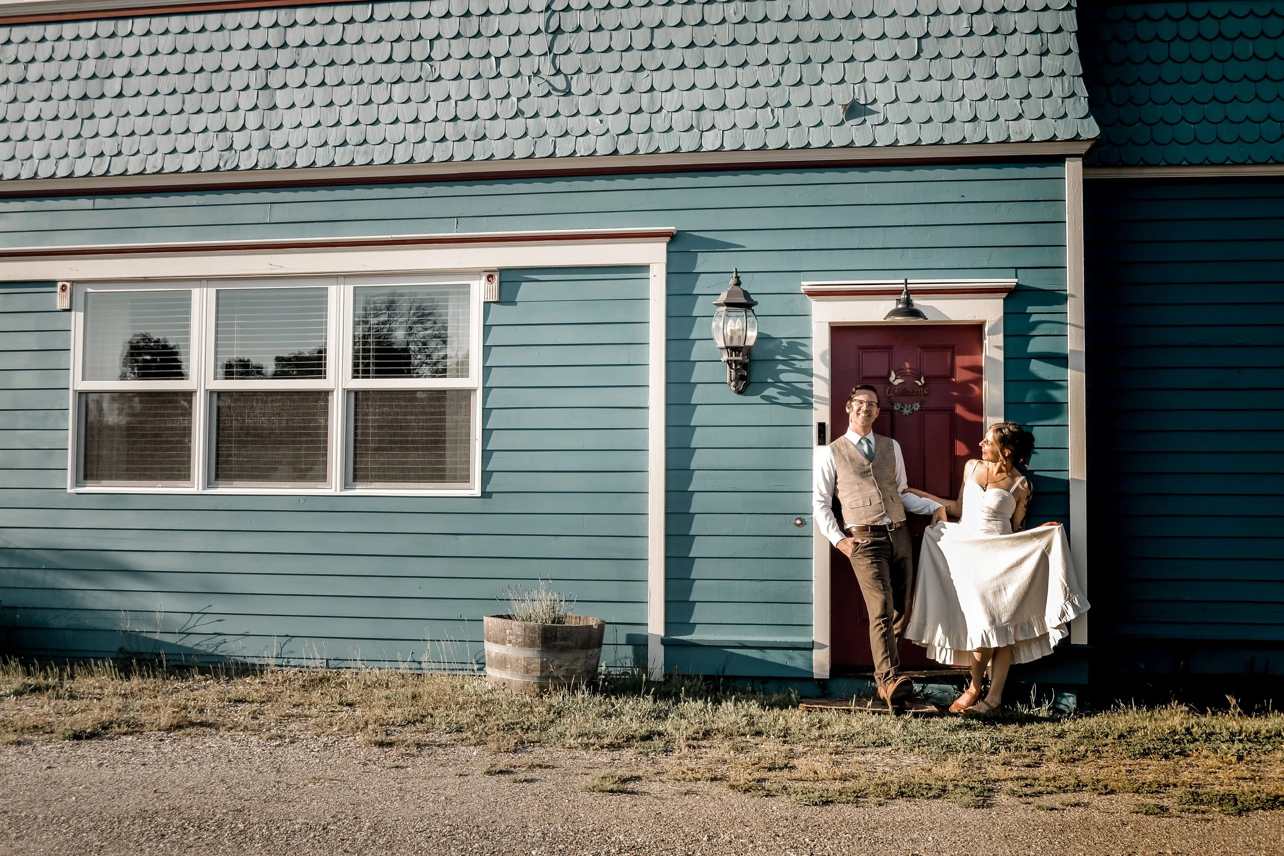 A man and woman standing outside a teal house with a red front door, during a wedding celebration. The woman is wearing a wedding dress, and the man is dressed in a vest and tie. They are smiling, with the woman holding her dress.