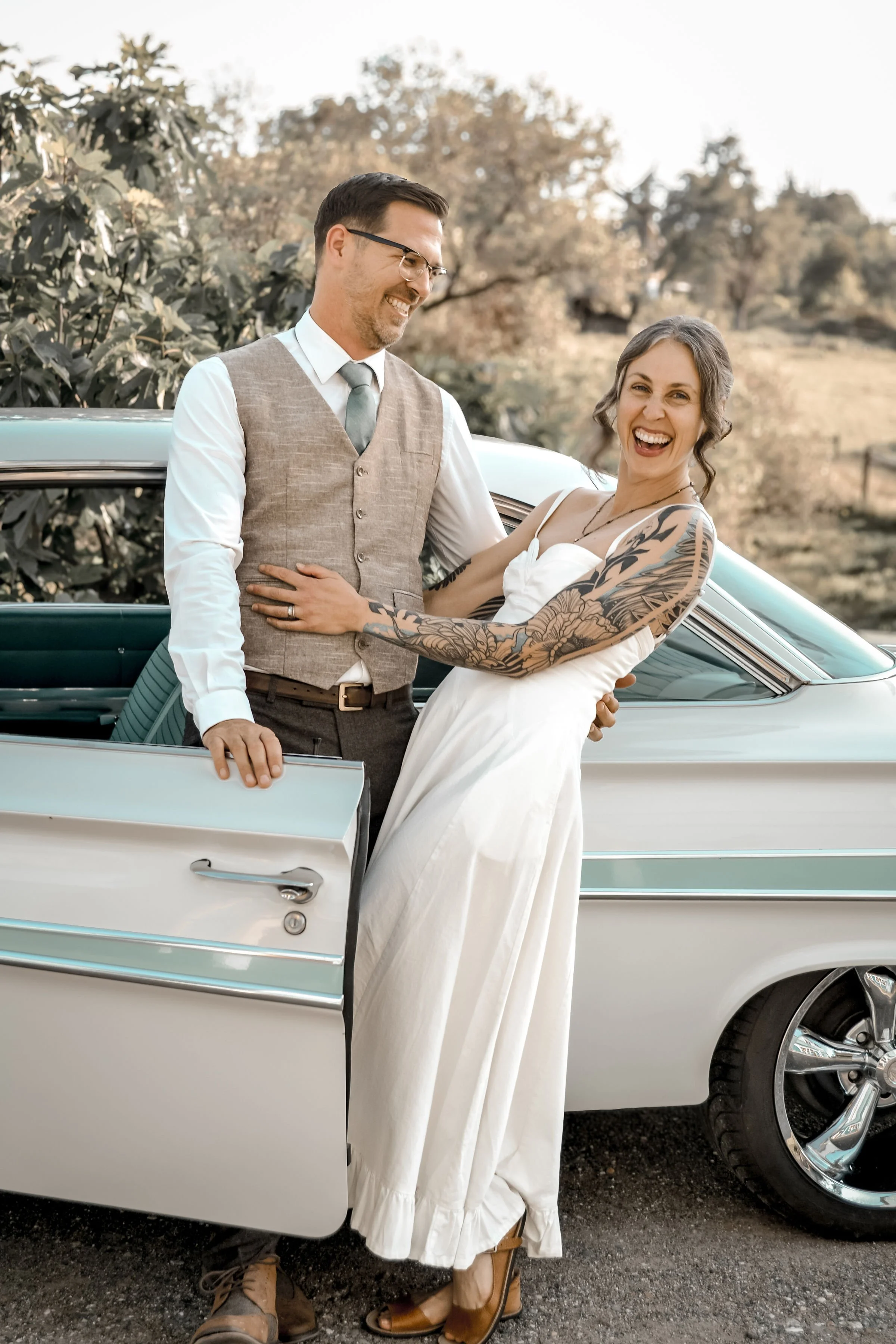 A woman in a white dress and tattoos on her arm is smiling and holding a man in a gray vest, white shirt, and glasses by a vintage car outdoors in a rural setting with trees in the background.