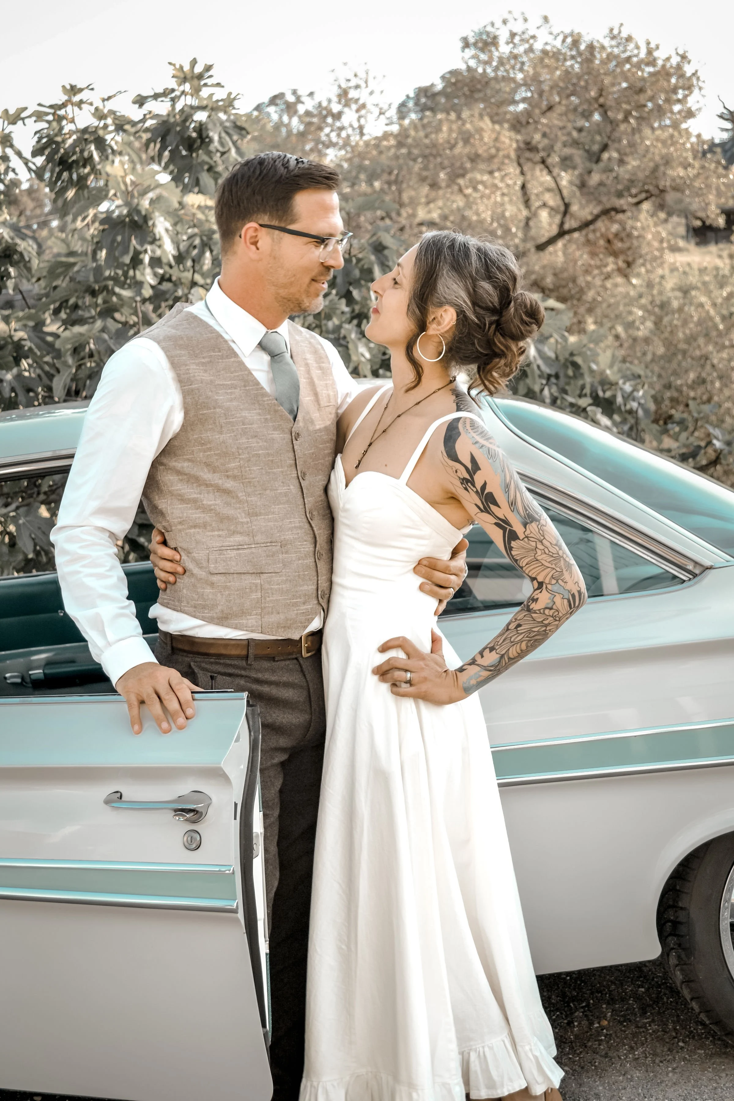 A couple stands close beside a vintage white car with a partially opened door, outdoors with trees in the background. The woman, wearing a white dress with thin straps and hoop earrings, has her hair styled in a bun and visible tattoos on her right a