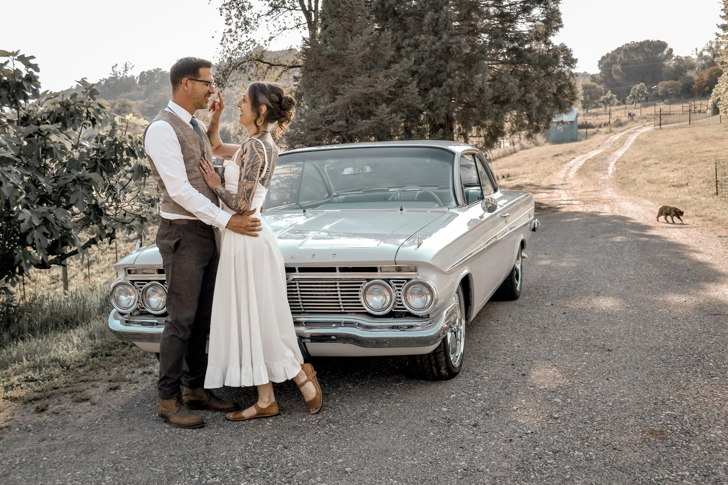 A couple stands close together near a vintage white car on a rural dirt road, with trees and hills in the background. The man wears glasses, a vest, and dress pants; the woman has tattoos, a white dress, and brown shoes.