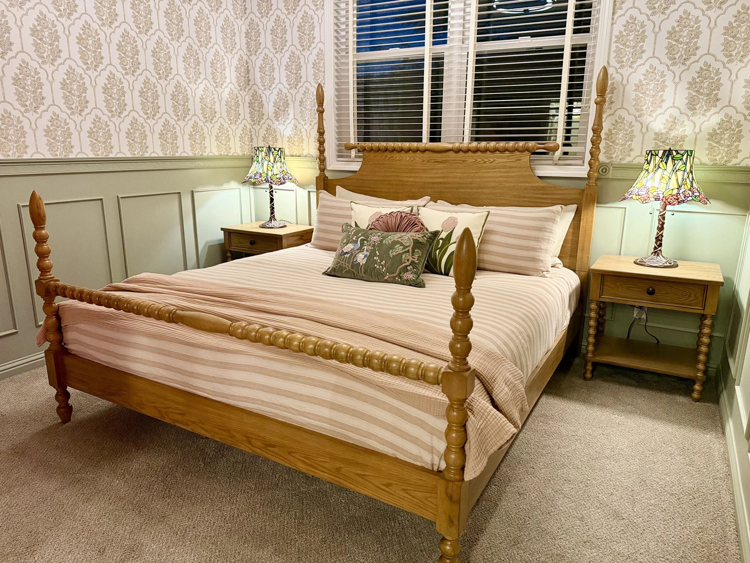 A cozy bedroom with a wooden four-poster bed, a headboard, and a floral decorative pillow, flanked by two nightstands with Tiffany-style lamps. The room has wallpaper with a floral pattern and light green wainscoting, with a large window behind the bed.