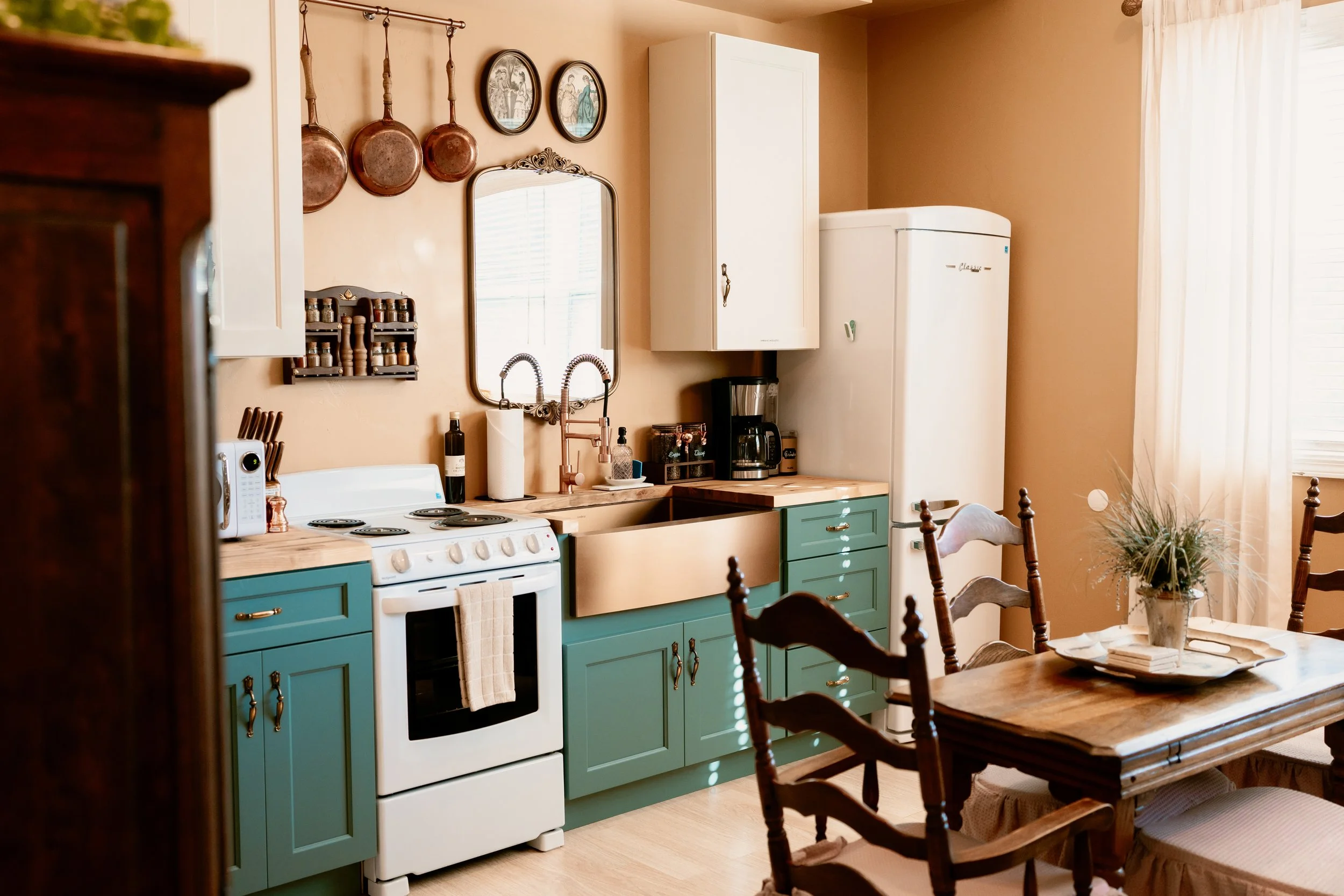 A cozy kitchen with teal cabinets, white appliances, including an oven and refrigerator, and a wooden dining table with chairs. Decor includes copper pans hanging on the wall, framed vintage artwork, and a small spice rack. A coffee maker and other small items are on the countertop, which is illuminated by natural light from a window with curtains.