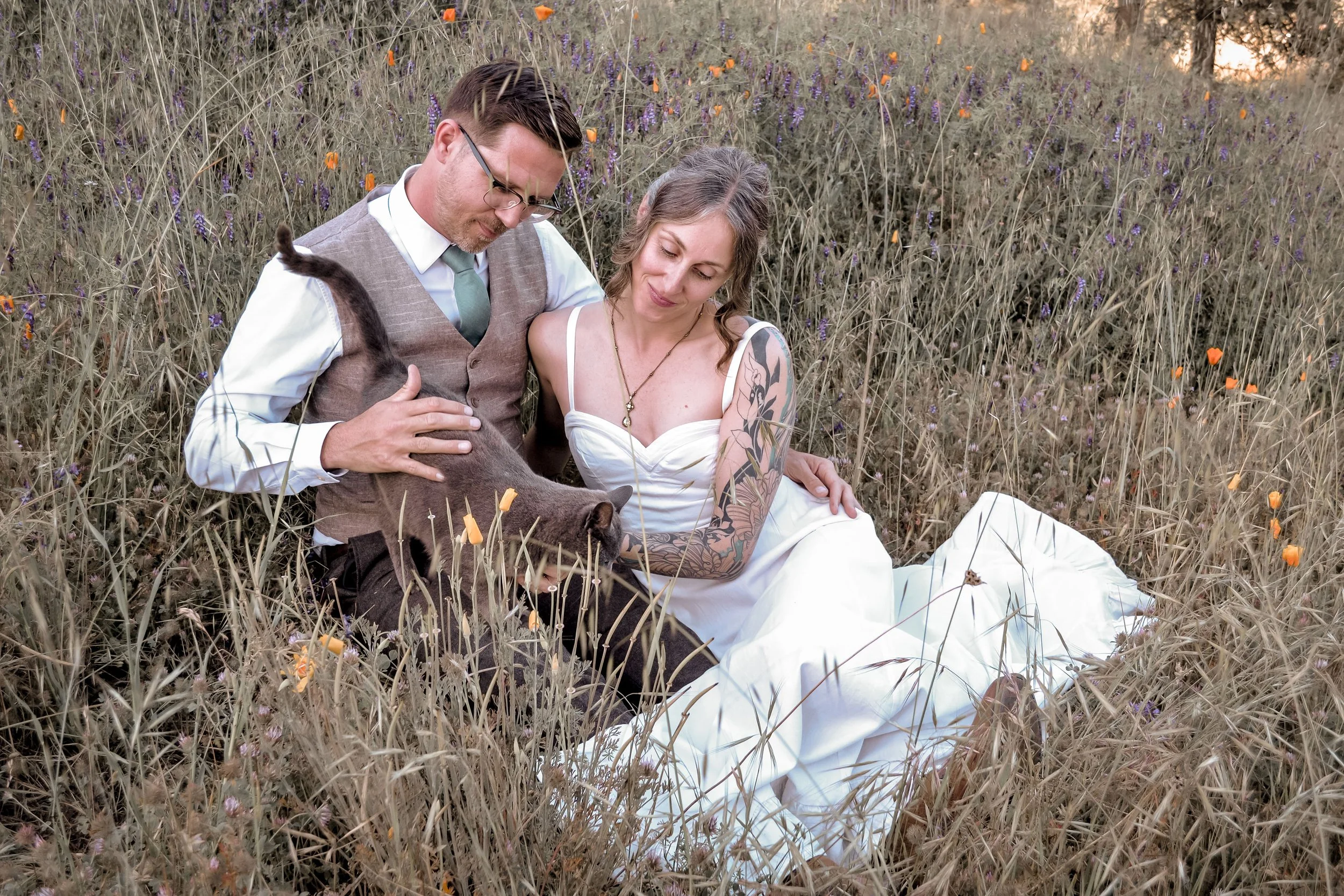 A man and woman sitting in a field of tall grasses and wildflowers, with purple and orange flowers, during sunset. The man wears a vest and glasses, and the woman has tattoos on her arm and is dressed in a white dress. They are close to each other, w