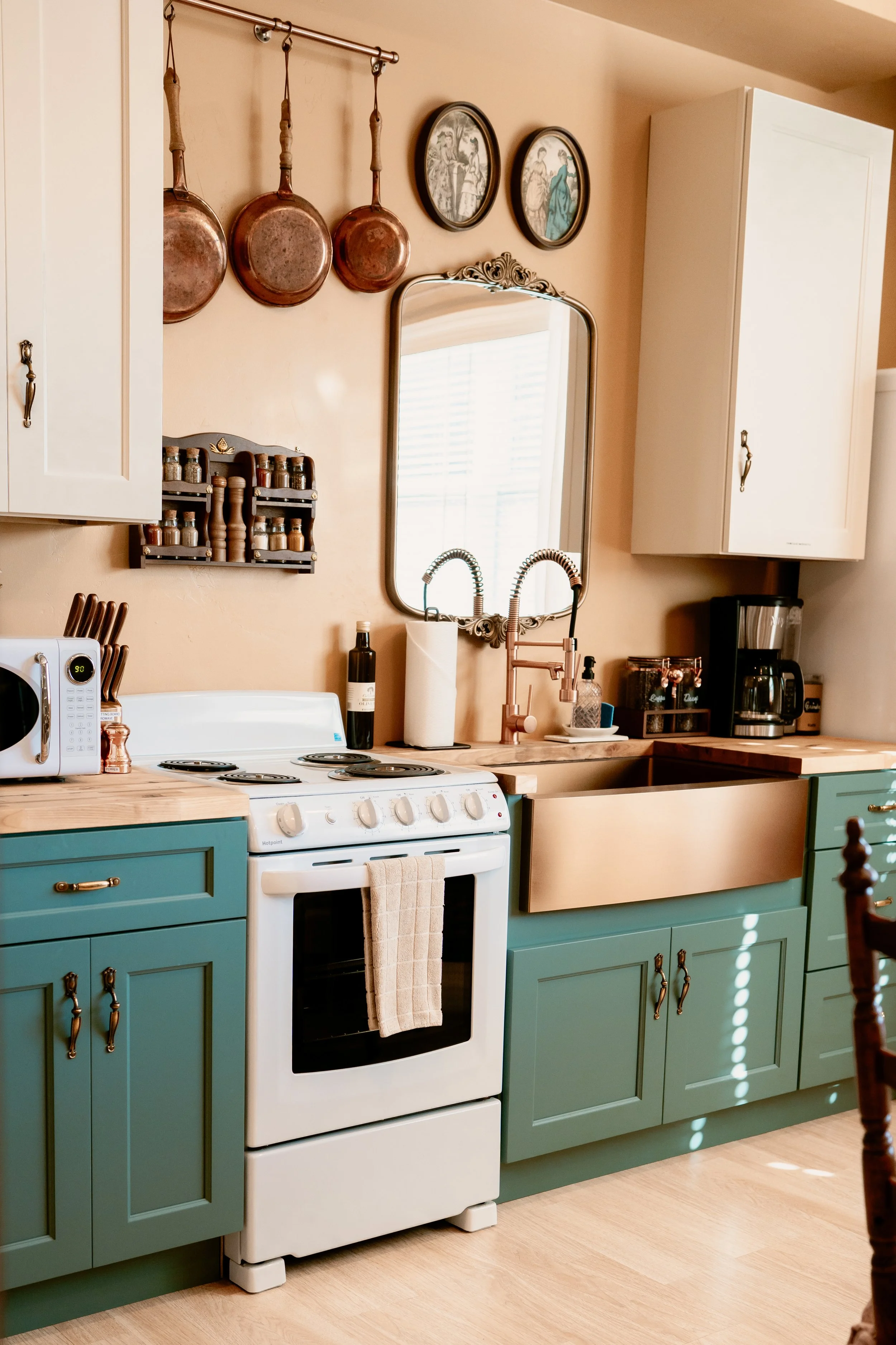 A kitchen corner with green cabinets and a white stove, copper sink, hanging copper pans, spice rack, mirror, and various kitchen appliances and decor.