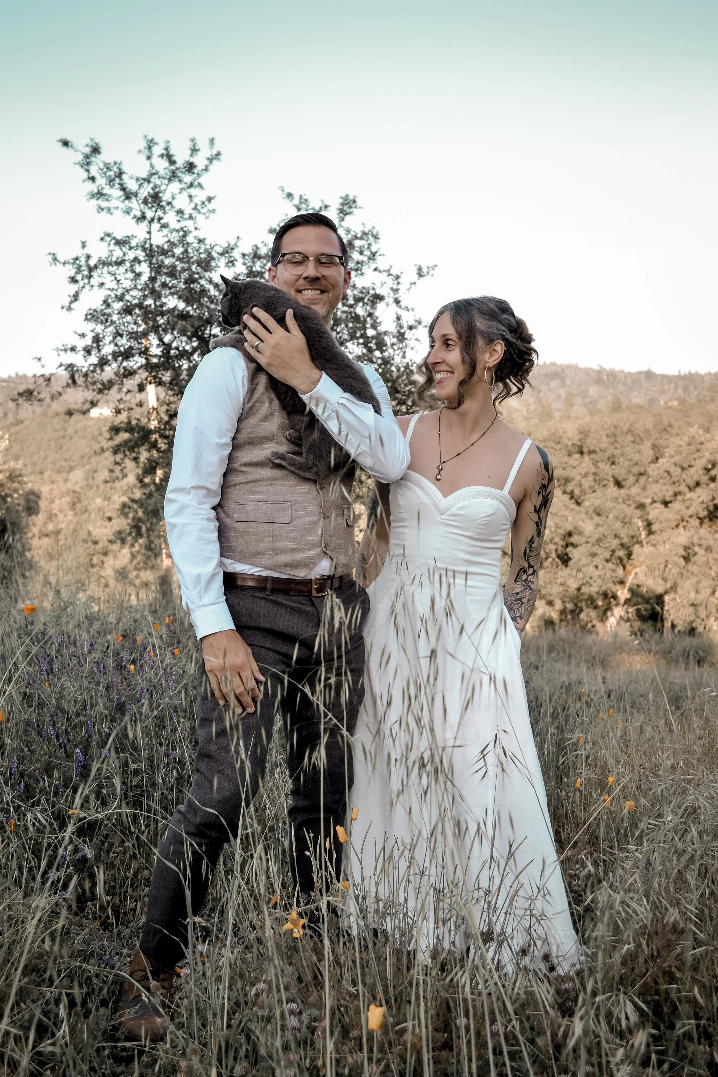A happy couple dressed in wedding attire is standing outdoors in a field of tall grass and wildflowers, with a tree and hills in the background. The man is holding a black cat, and the woman is smiling at him.