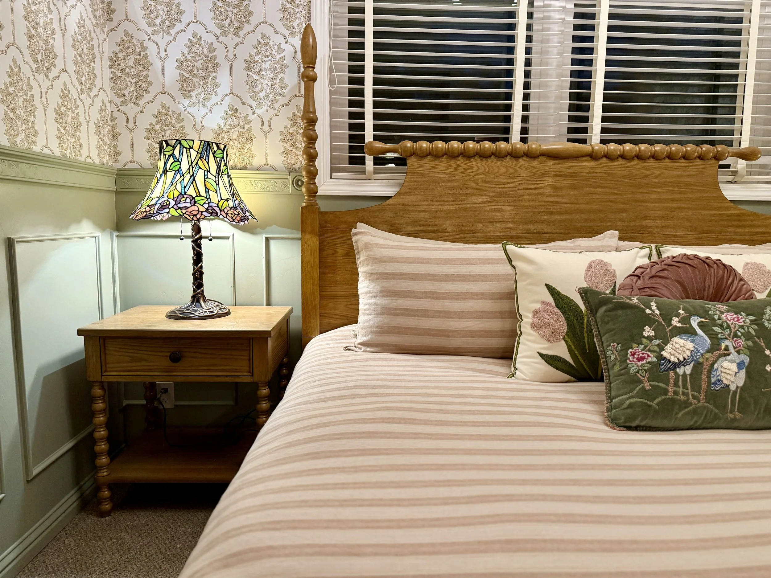 A bedroom scene featuring a wooden bed with pillows, a Tiffany-style stained glass lamp on a wooden nightstand, and a window with blinds. The wall has patterned wallpaper and wood paneling.