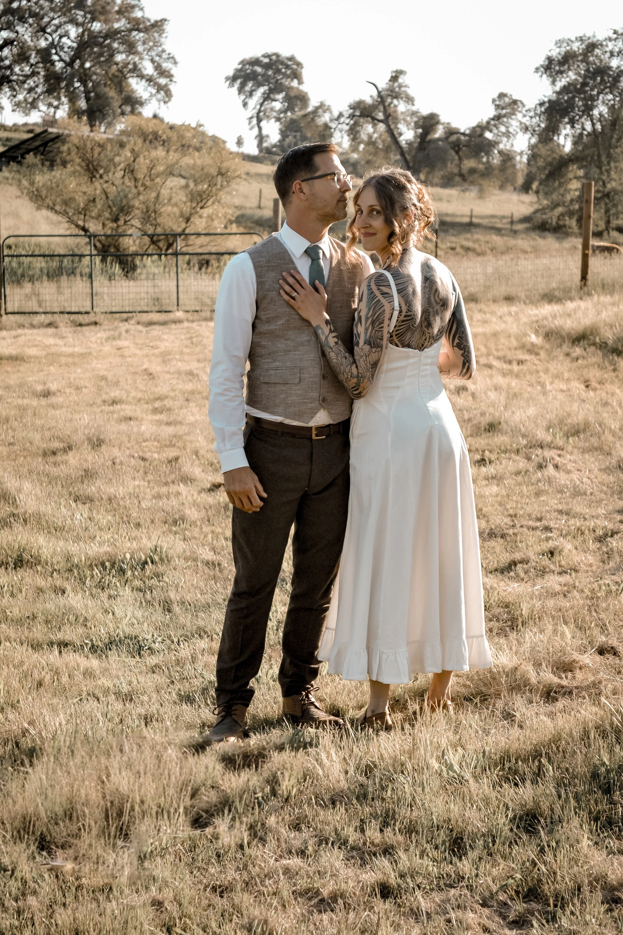A couple standing in a grassy field with trees in the background, dressed in vintage or rustic-style clothing. The woman is smiling, touching the man's chest, and wearing a white dress with a tattoo sleeve on her arm. The man is wearing glasses, a ve