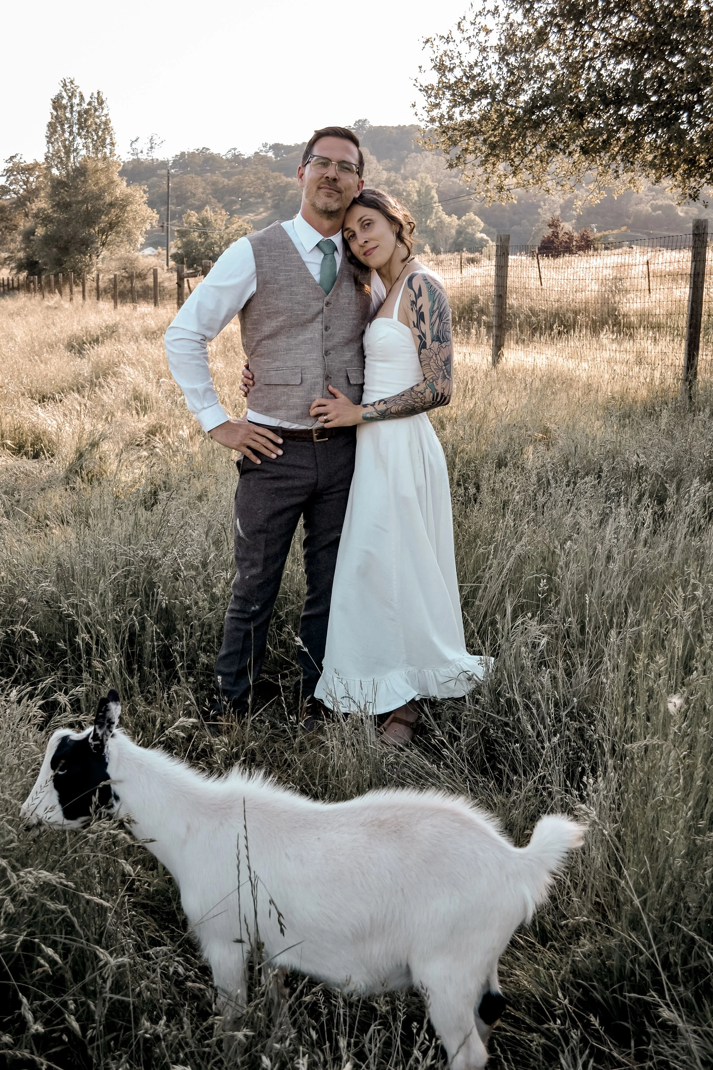A couple standing close together in a field, with a white and black dog in the foreground. The man is wearing glasses, a white shirt, and a gray vest, and the woman is in a white dress with tattoos on her arms. They are outdoors with trees and hills 