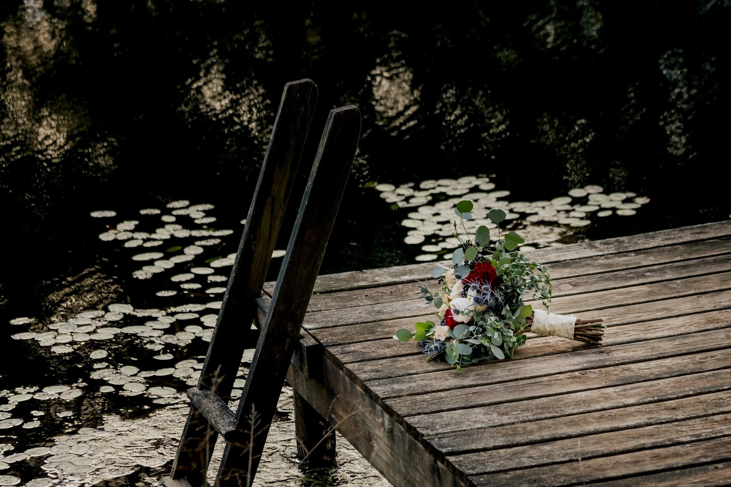 A bouquet of flowers resting on a weathered wooden dock beside a dark pond with lily pads.