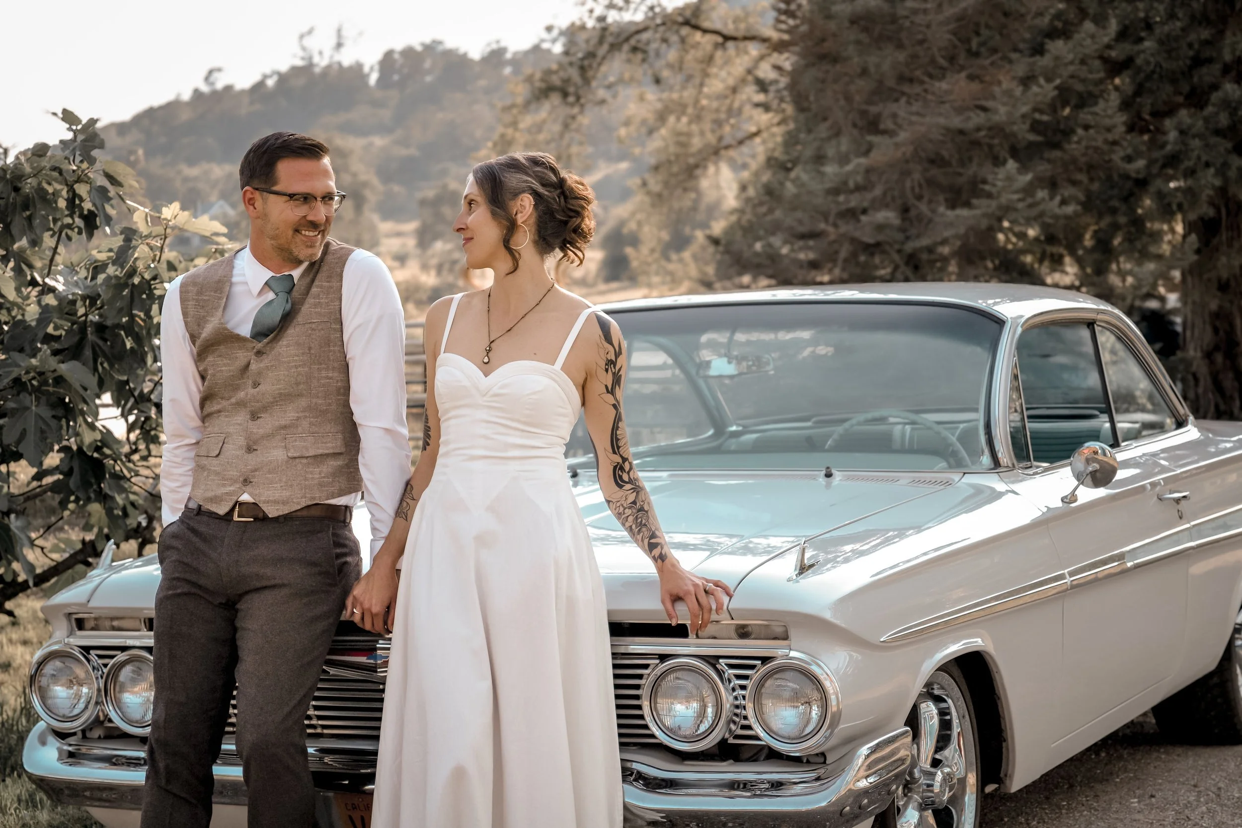 A smiling man and woman holding hands, standing in front of a classic white convertible car outdoors, with hills and trees in the background. The woman is wearing a white dress and the man is dressed in a vest, shirt, and tie.