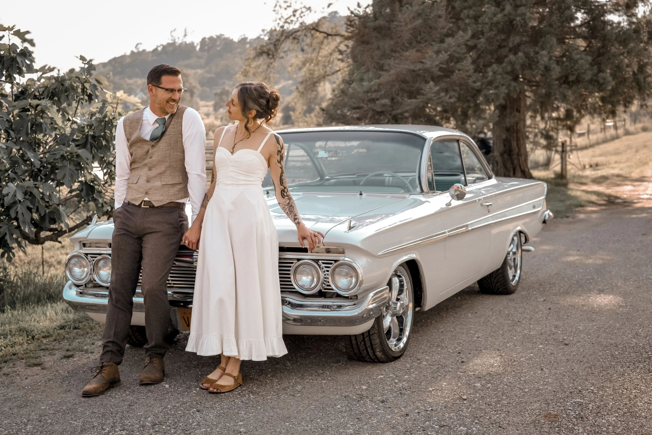 A man and woman stand together in front of a white vintage car outdoors. The woman wears a white dress and the man wears a vest and tie. They are smiling at each other with trees and hills in the background.