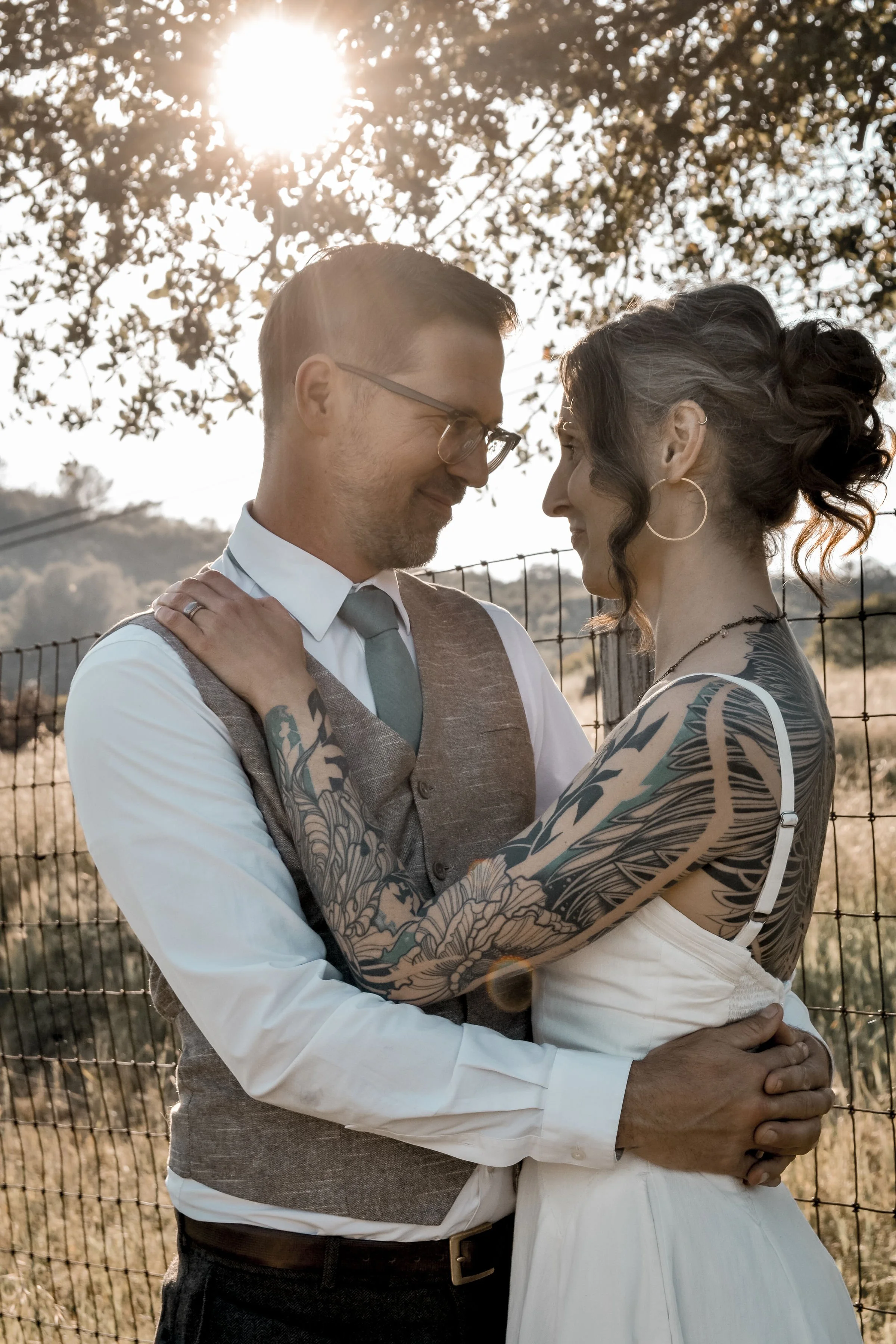 A couple standing close together outdoors, embracing each other with their faces touching, in front of a wire fence with trees and the sun shining behind them.