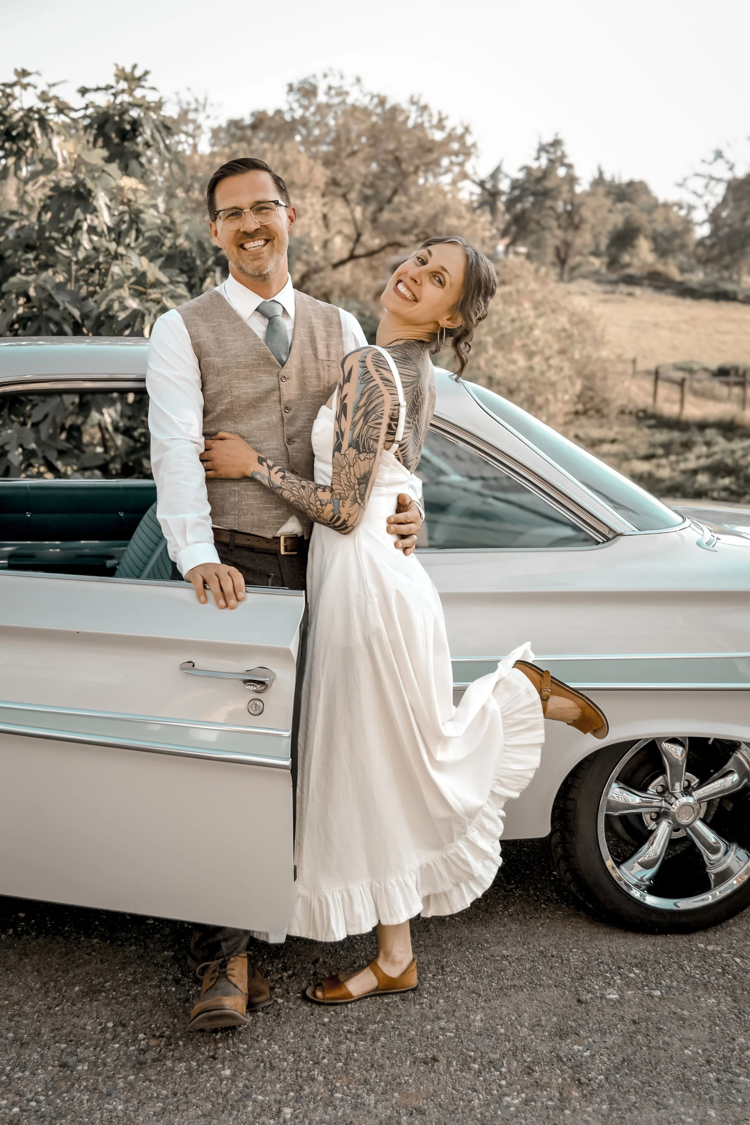 A happy couple standing next to a vintage car outdoors, with trees and a clear sky in the background. The woman has tattoos and is wearing a white dress with brown sandals, while the man is dressed in a vest, shirt, tie, and glasses.