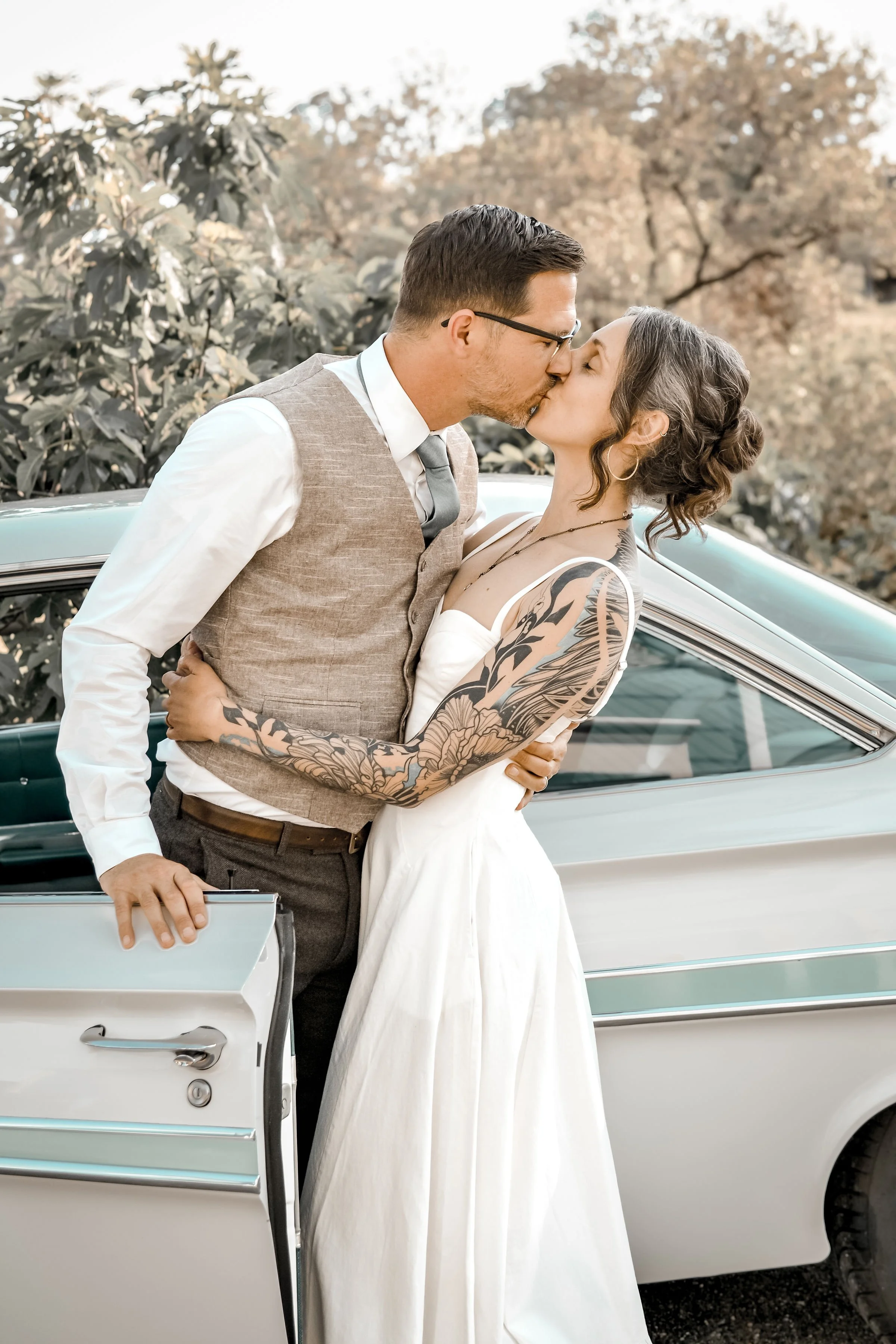 A man and woman sharing a kiss in front of a vintage car outdoors, with trees in the background.