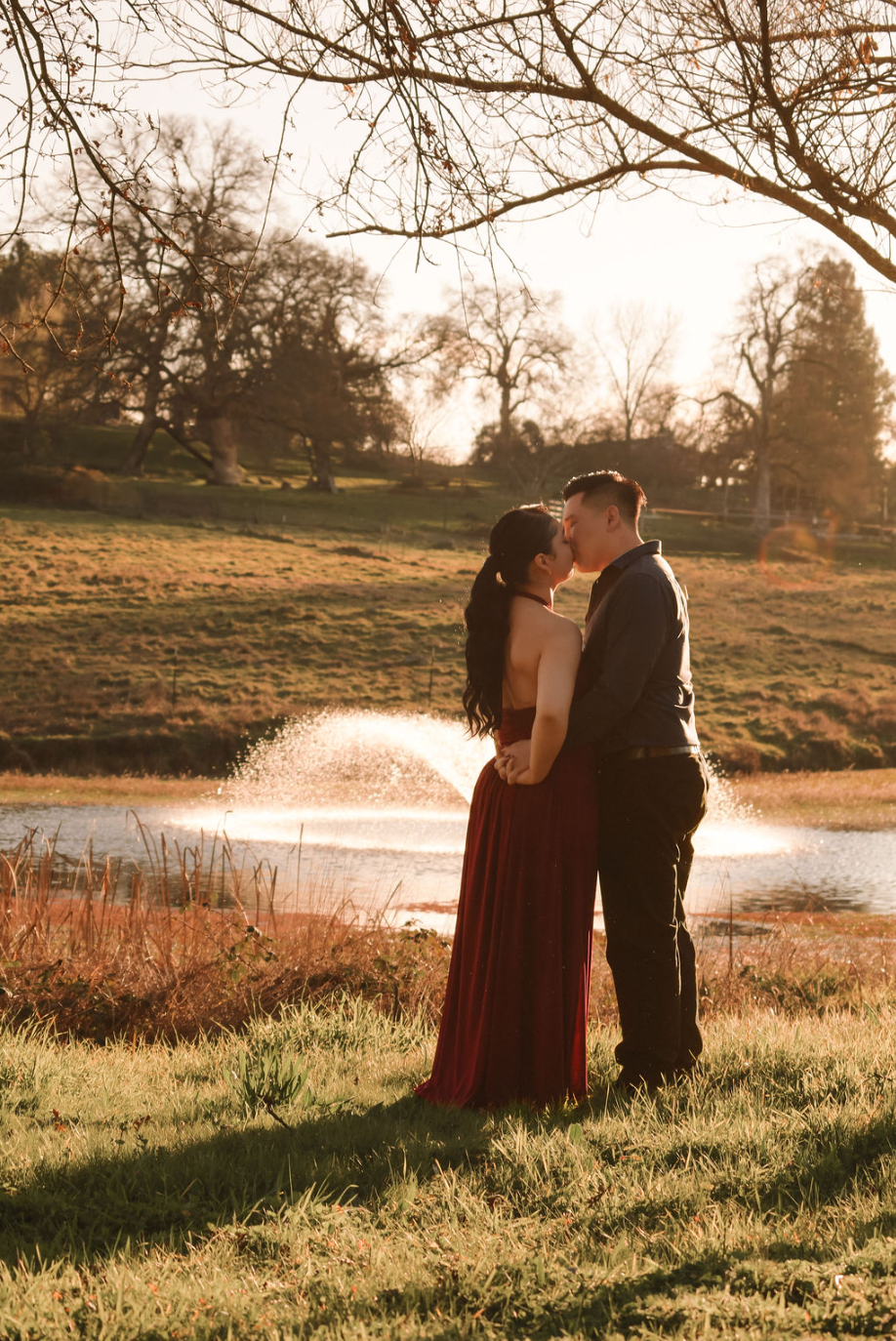 A couple kissing outdoors near a pond during sunset, with trees and grass in the background.