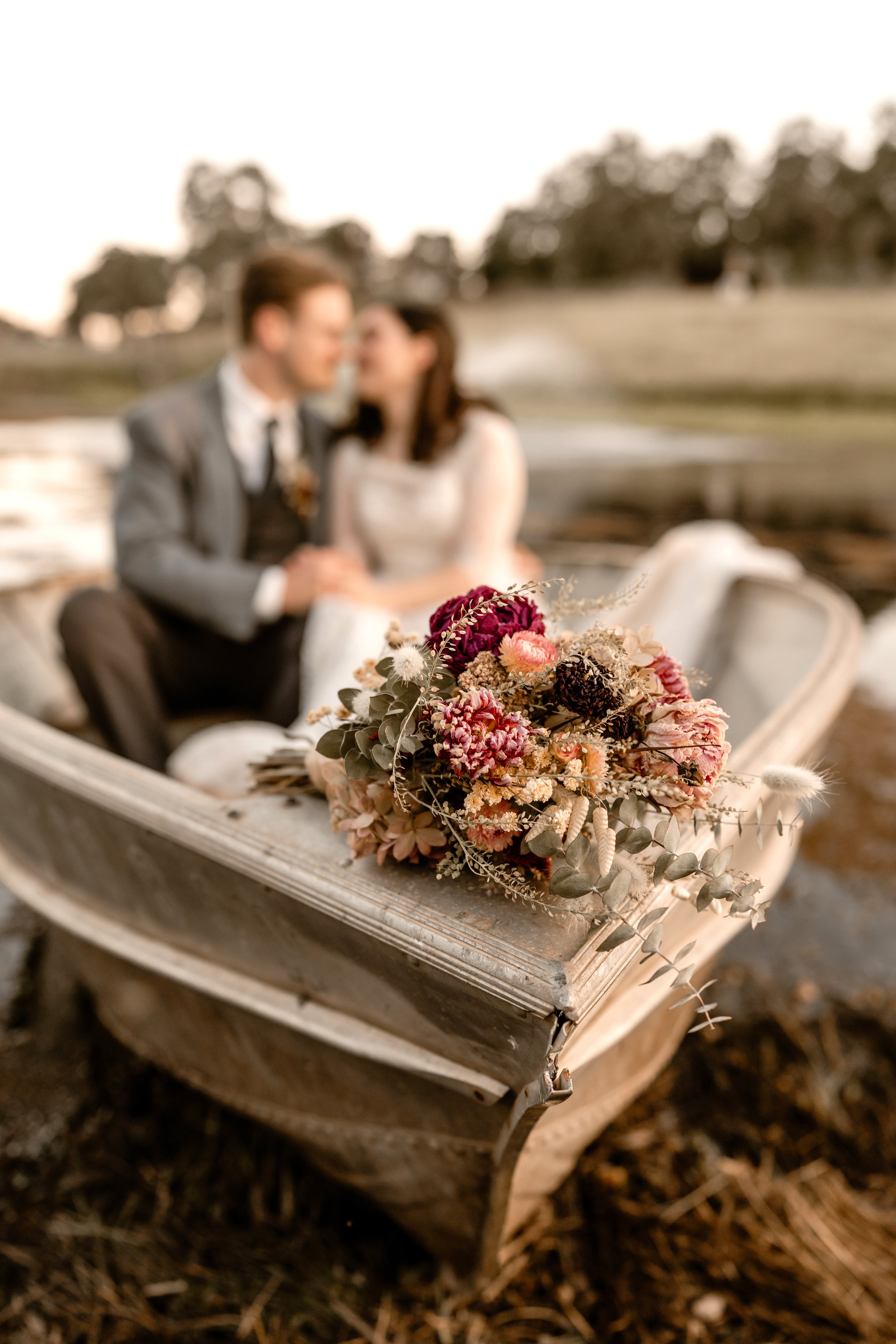 A bouquet of dried flowers with pink, purple, and beige colors resting on the edge of an old, white wooden rowboat with chipped paint, on dry ground with the blurred background of a park or field.