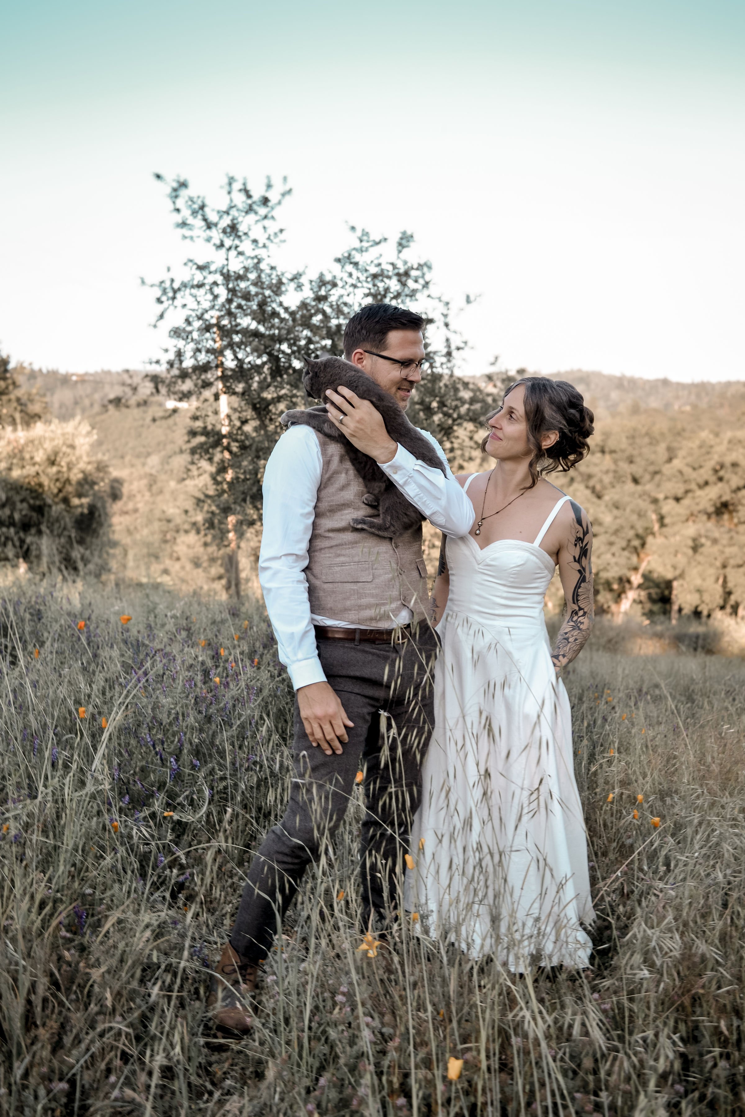 A couple standing in a field with wildflowers, the man holding a gray cat and looking at the woman, the woman gazing at the man, the backdrop of trees and a clear sky.