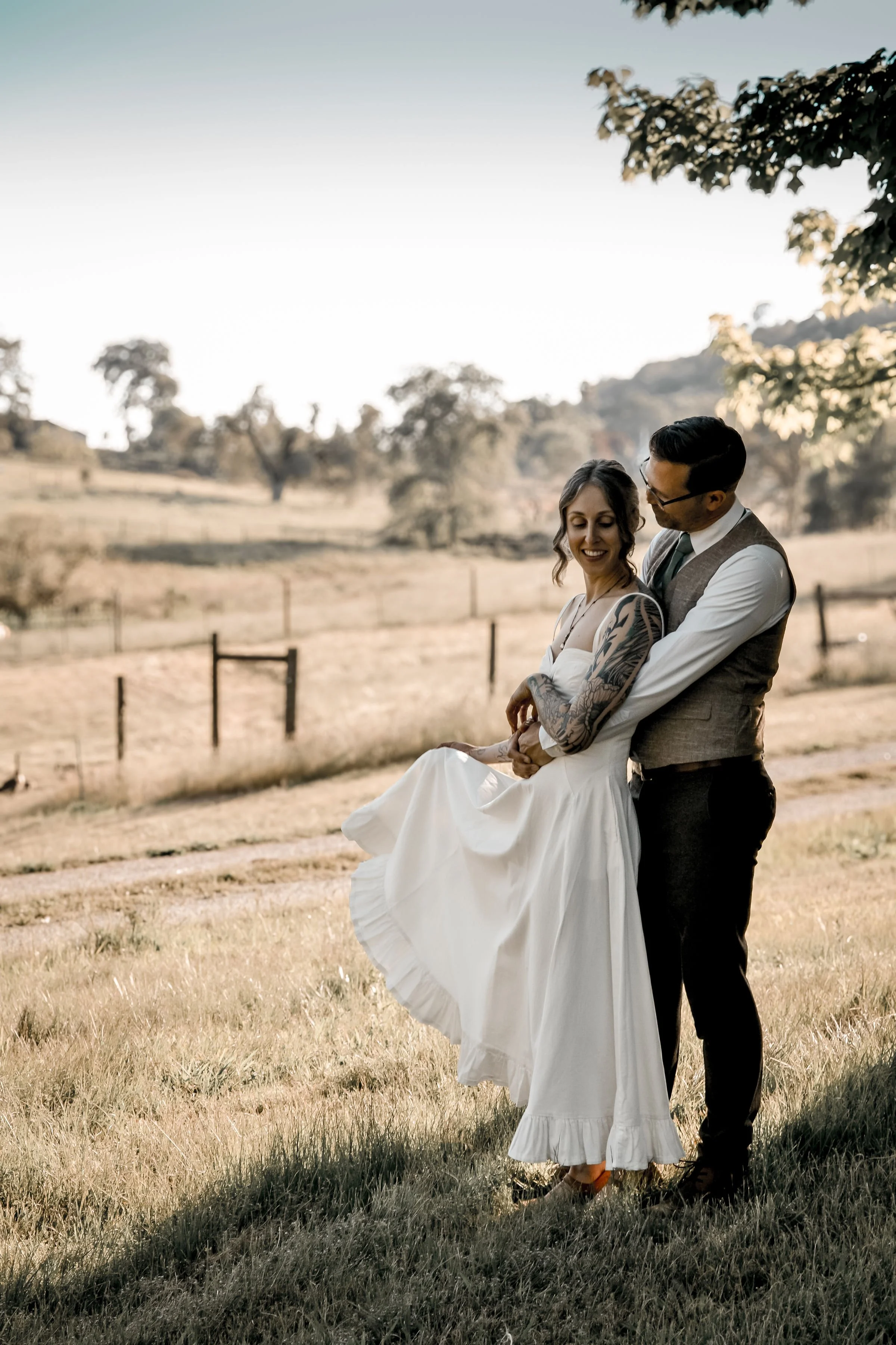 A couple dressed in wedding attire standing close together in an outdoor grassy field, smiling and sharing an intimate moment, with a scenic background of trees and distant hills during daylight.