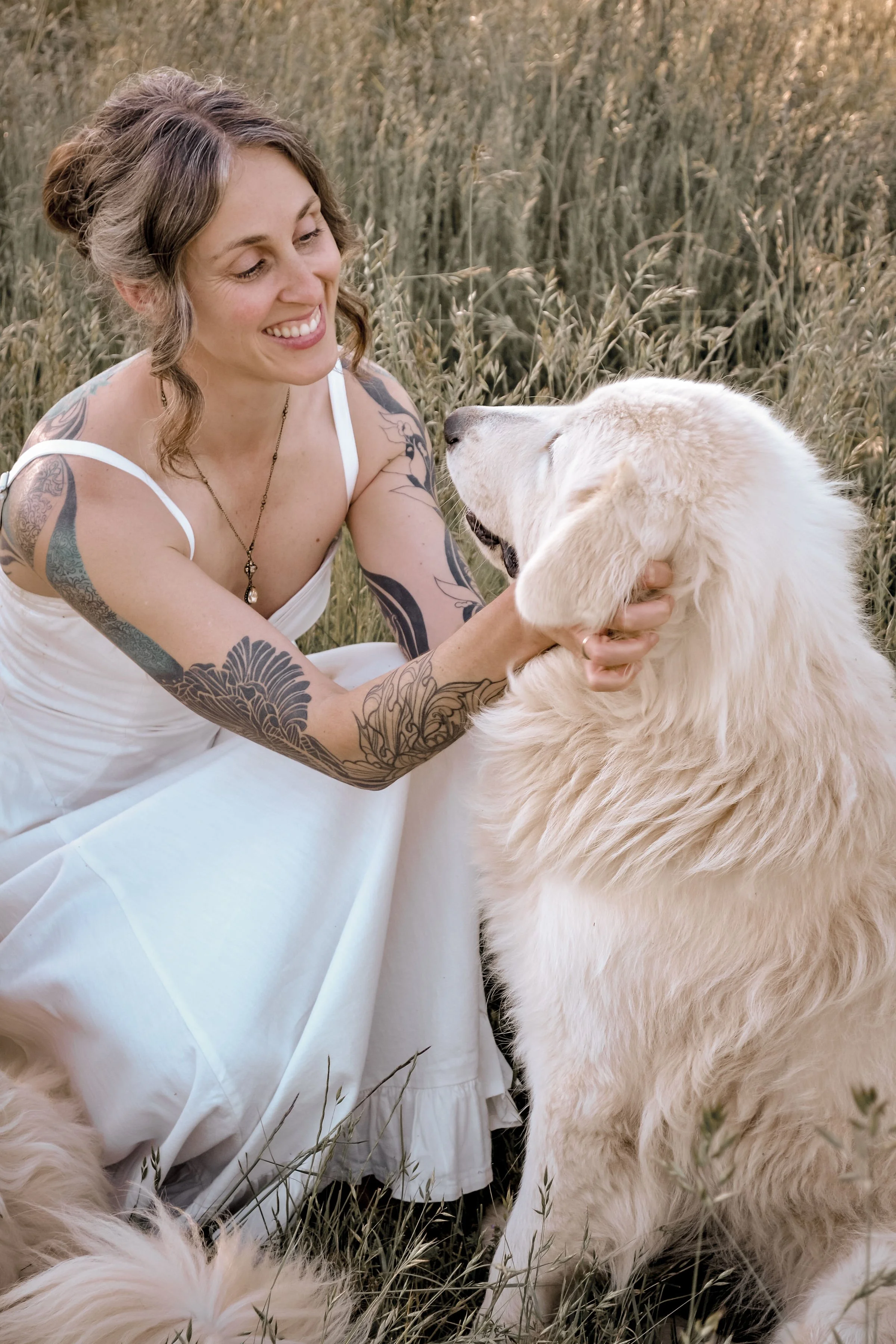 A woman with tattoos smiling while holding a golden retriever dog in a field of tall grass.