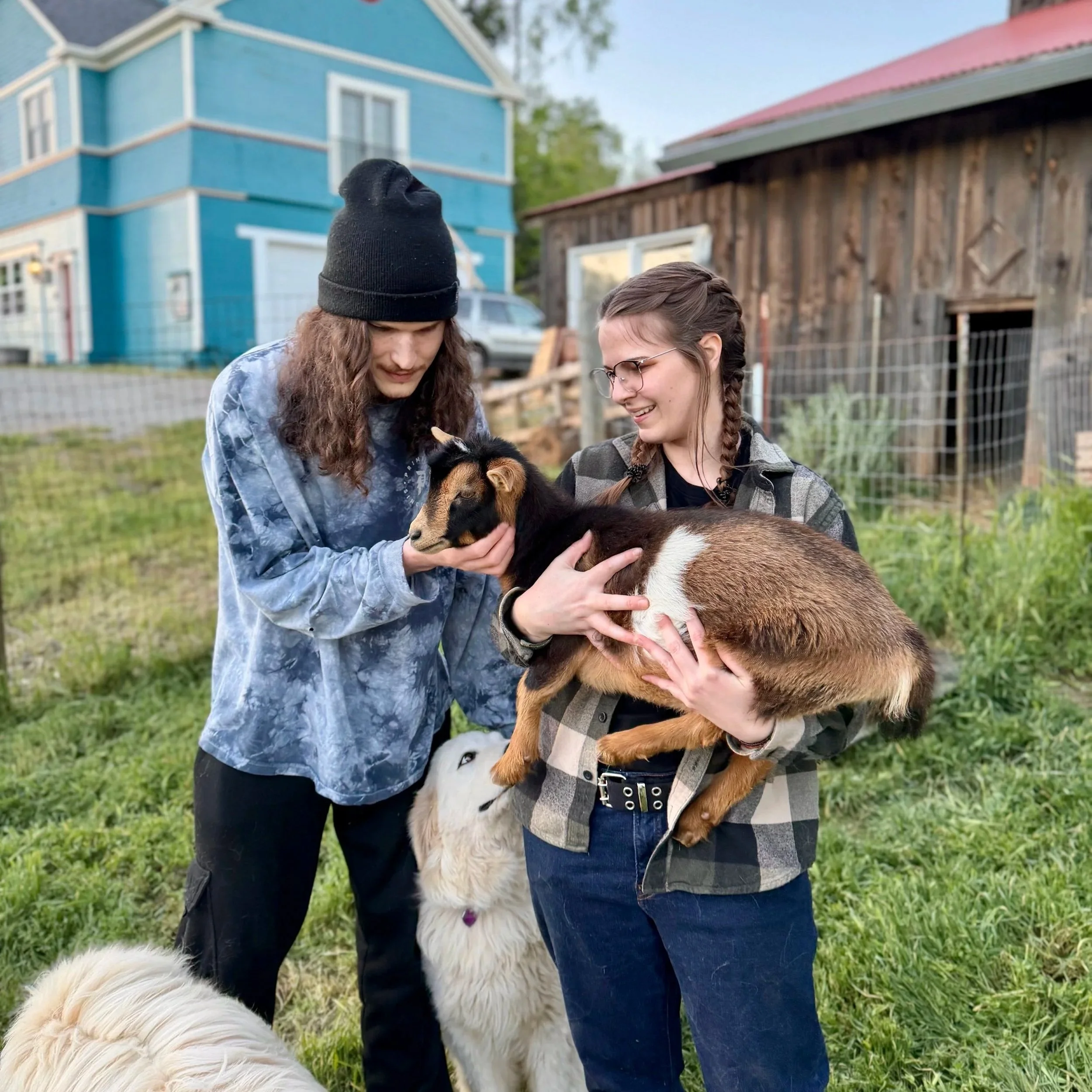 Two young women outdoors with dogs, one holding a large fluffy dog and the other looking at it, background of a blue house and a wooden barn.