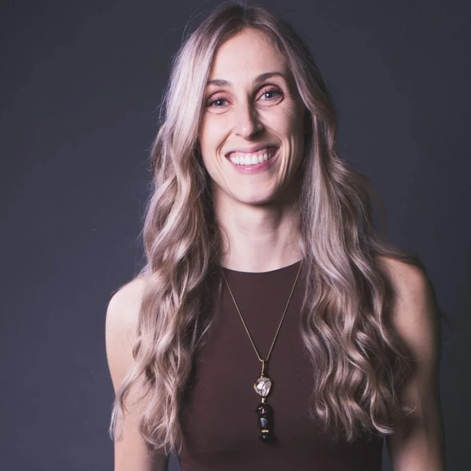 Smiling woman with long, wavy blonde hair, wearing a sleeveless maroon top and a necklace with a black pendant, against a dark gray background.