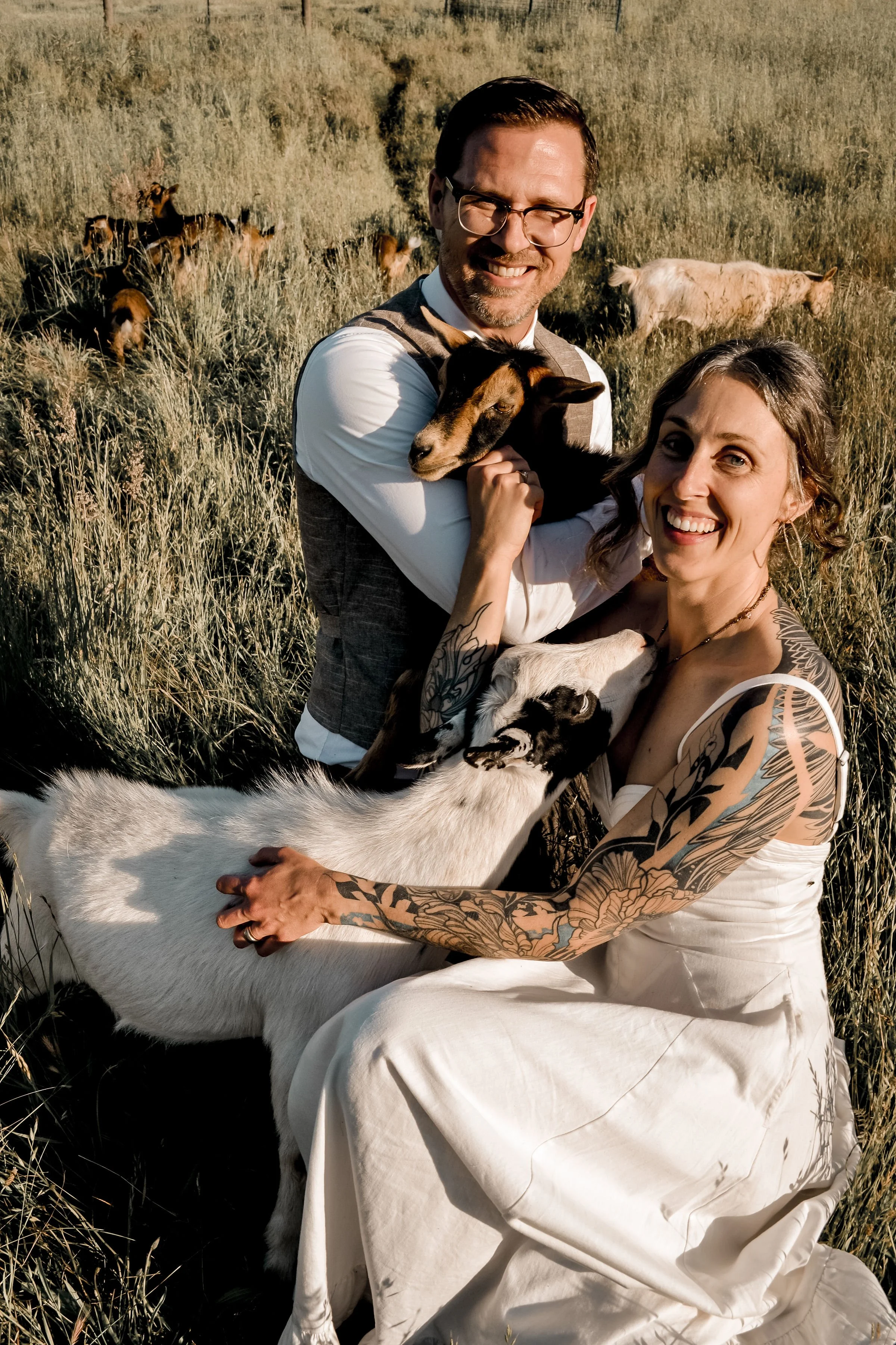 A happy man and woman pose in a field with goats and sheep, with the man holding a goat and the woman holding a sheep, all smiling.