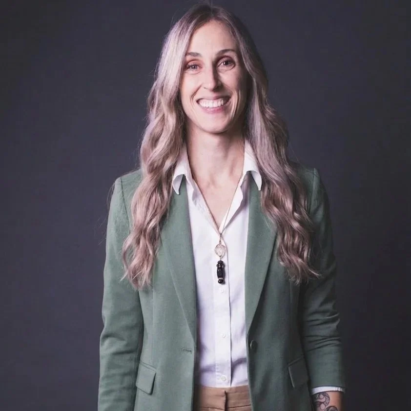 Smiling woman with long wavy hair wearing a green blazer, white shirt, and necklace against a dark background.