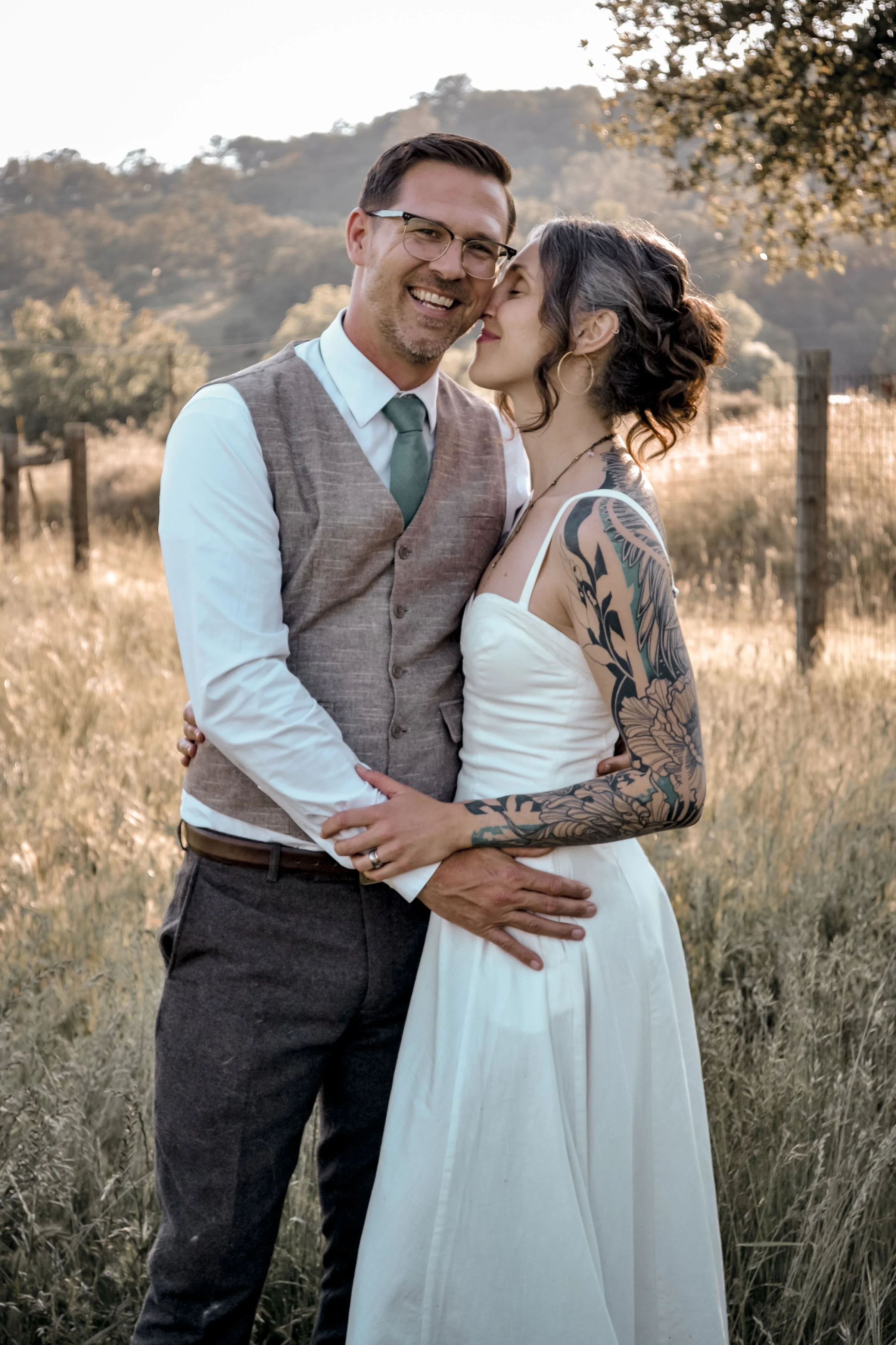 A couple standing in a grassy field with hills and trees in the background during sunset, close to each other, smiling and sharing an intimate moment.