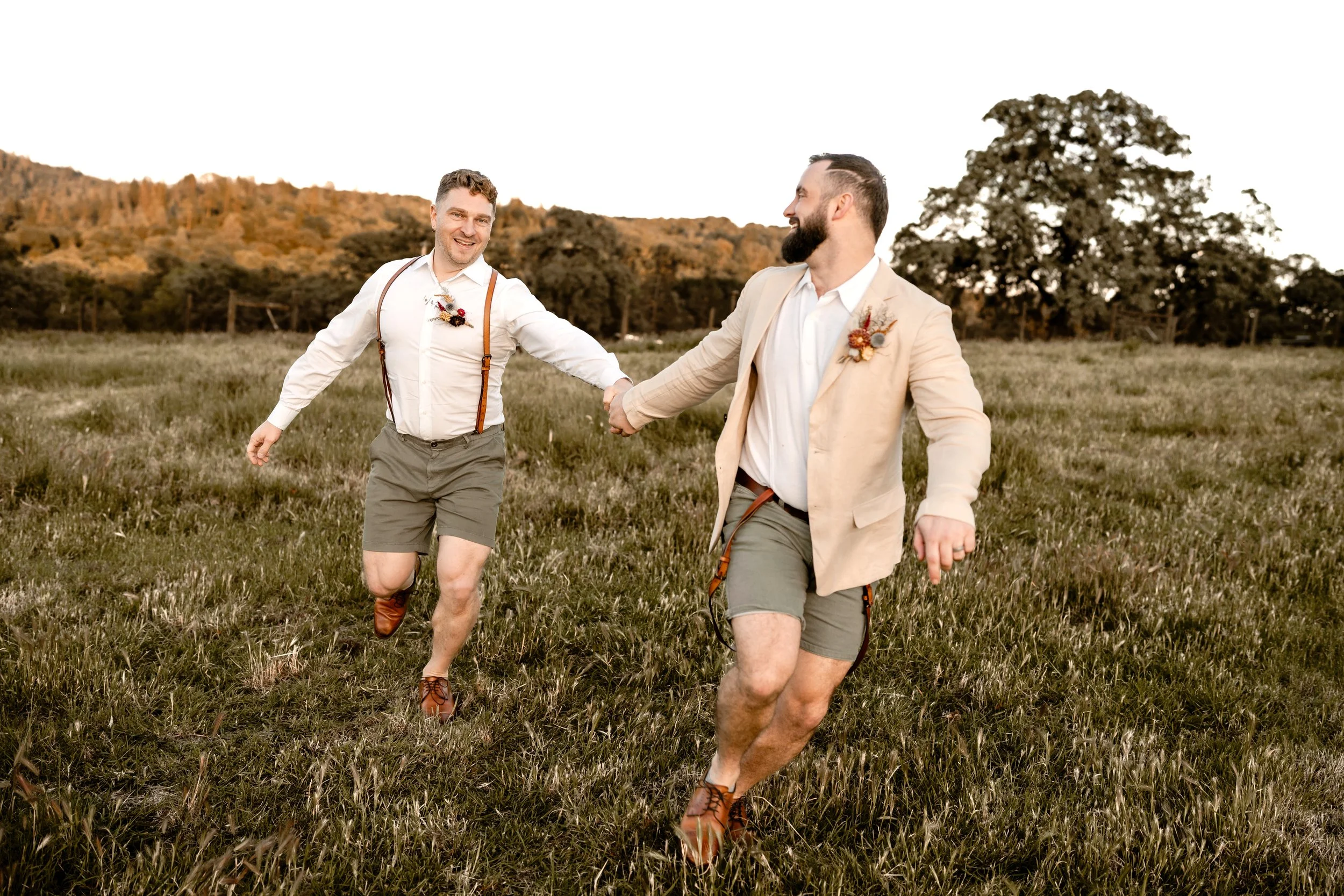 Two men are walking hand in hand through a grassy field with a backdrop of hills and trees. They are smiling and dressed in semi-formal attire with boutonnières on their jackets, indicating a wedding or celebration.