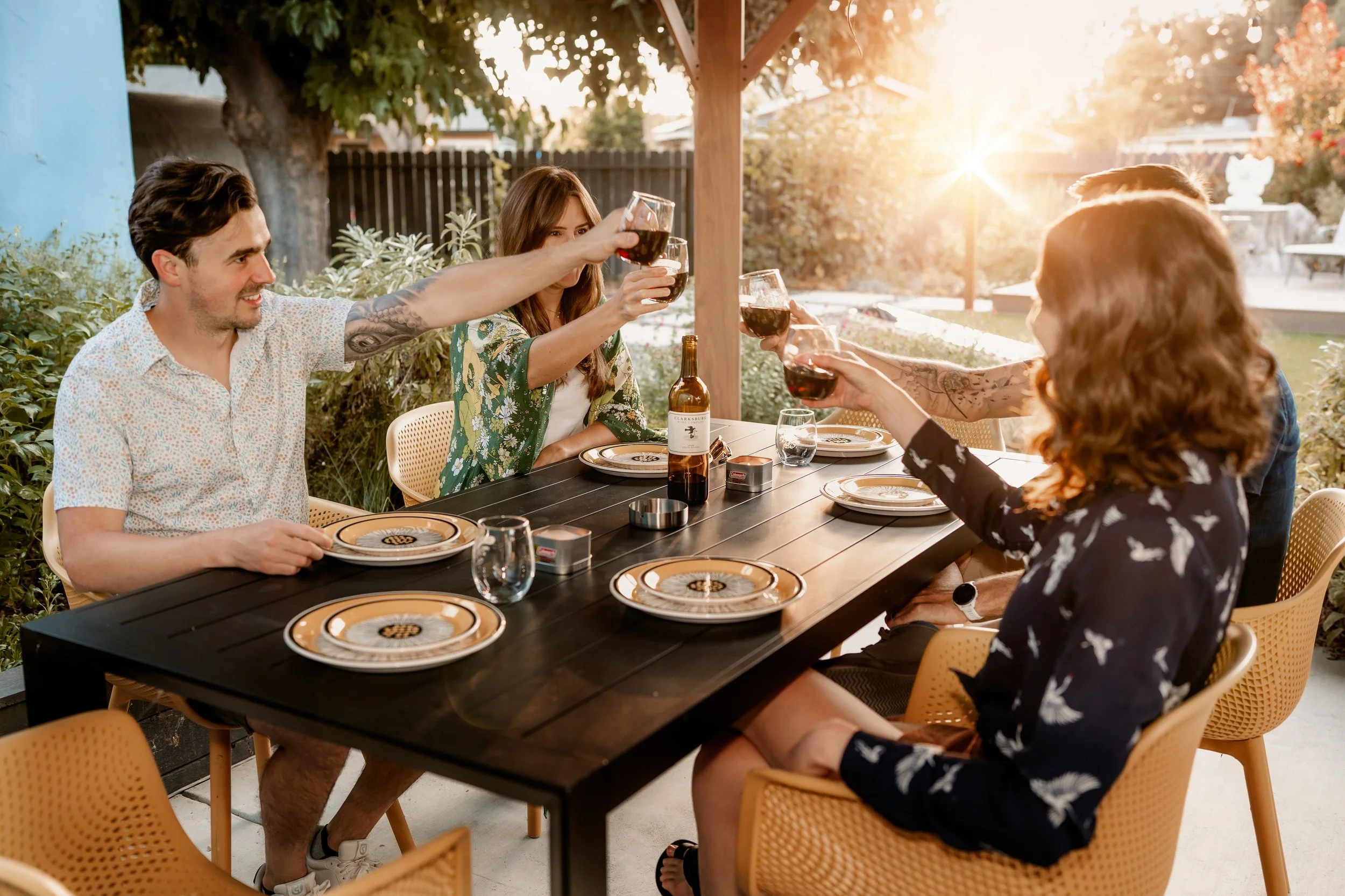 A group of friends sitting around a black outdoor table under a wooden gazebo, raising glasses of red wine in a toast, enjoying a meal outdoors during sunset.