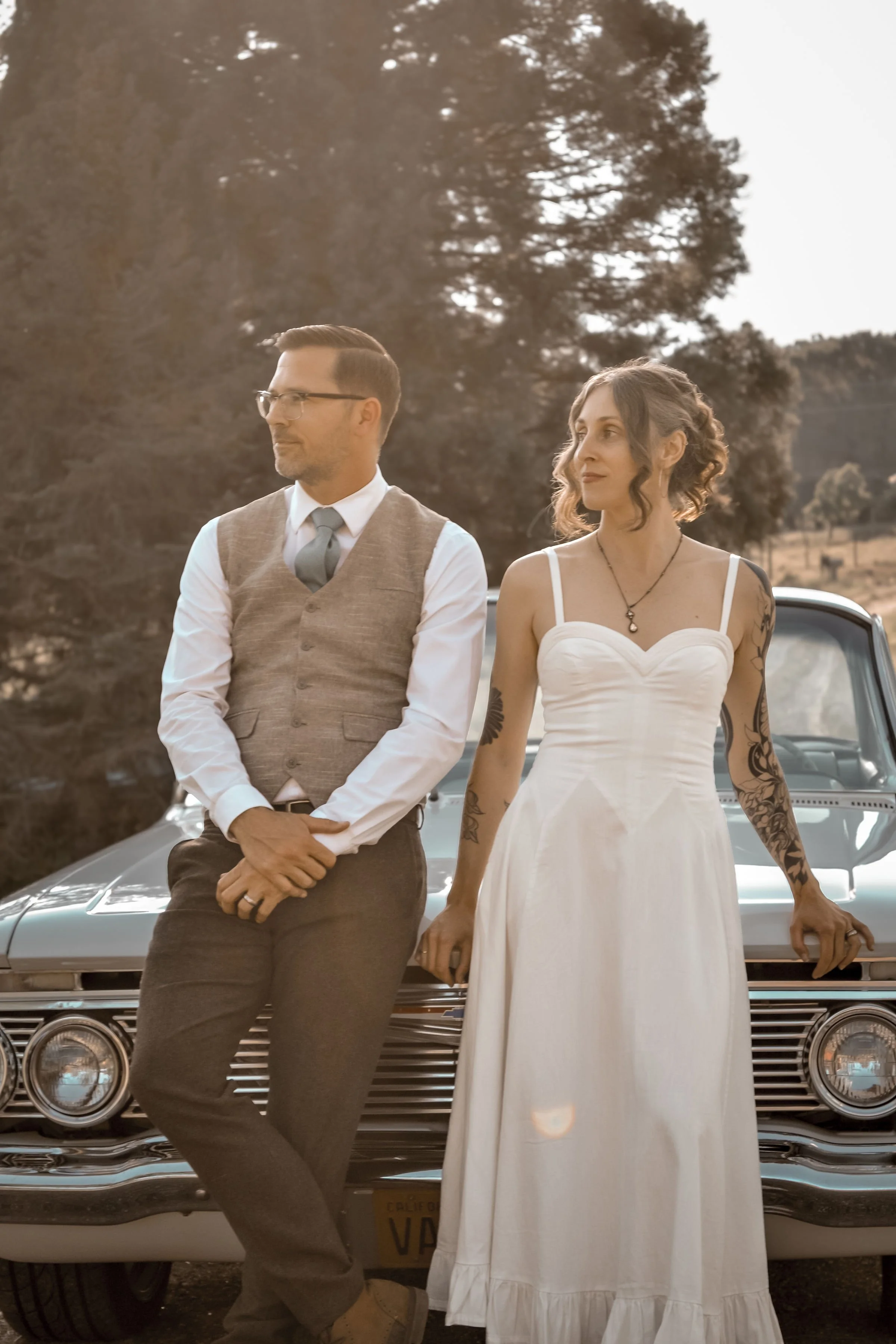 A man in a gray vest and tie standing with his hands clasped in front of him, and a woman in a white dress with tattoos on her arms standing with her hand on the car, with a vintage car and trees in the background during sunset.