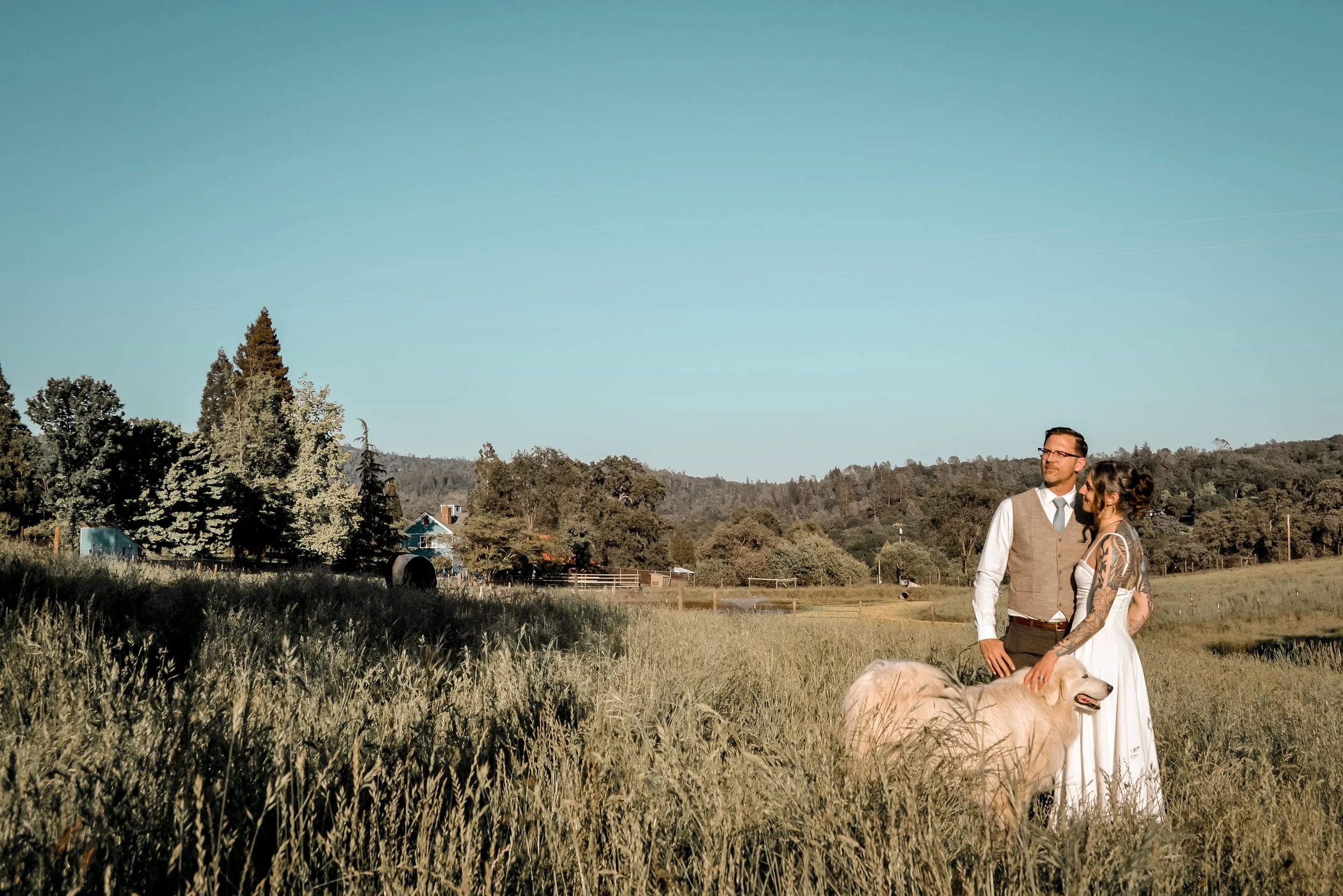A couple with tattoos and glasses sitting outdoors on a grassy field with a white dog, with trees, hills, and blue sky in the background.