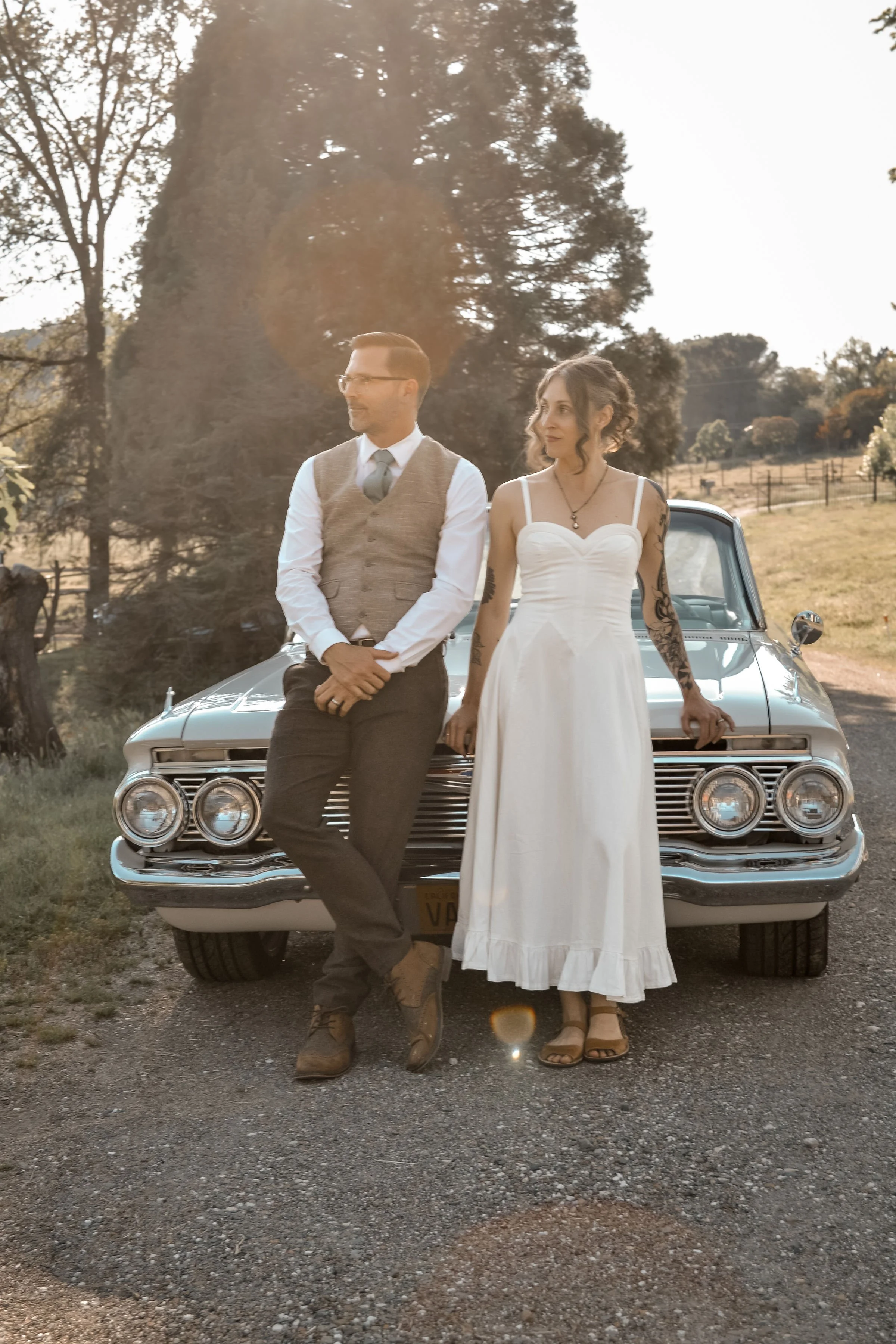 A man and woman standing in front of a vintage car outdoors during golden hour. The man is wearing a brown vest, white shirt, and tie, and the woman is wearing a white dress with tattoos on her arms.