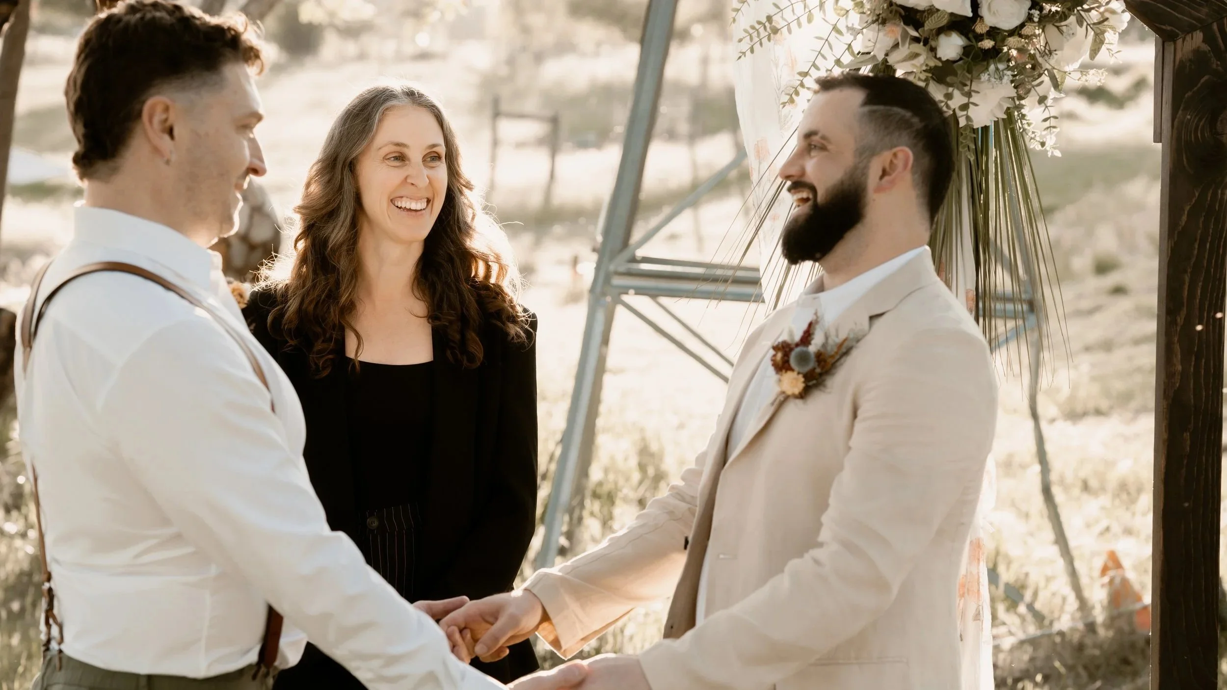 A couple getting married outdoors, holding hands and facing each other, with an officiant and floral arrangement in the background.