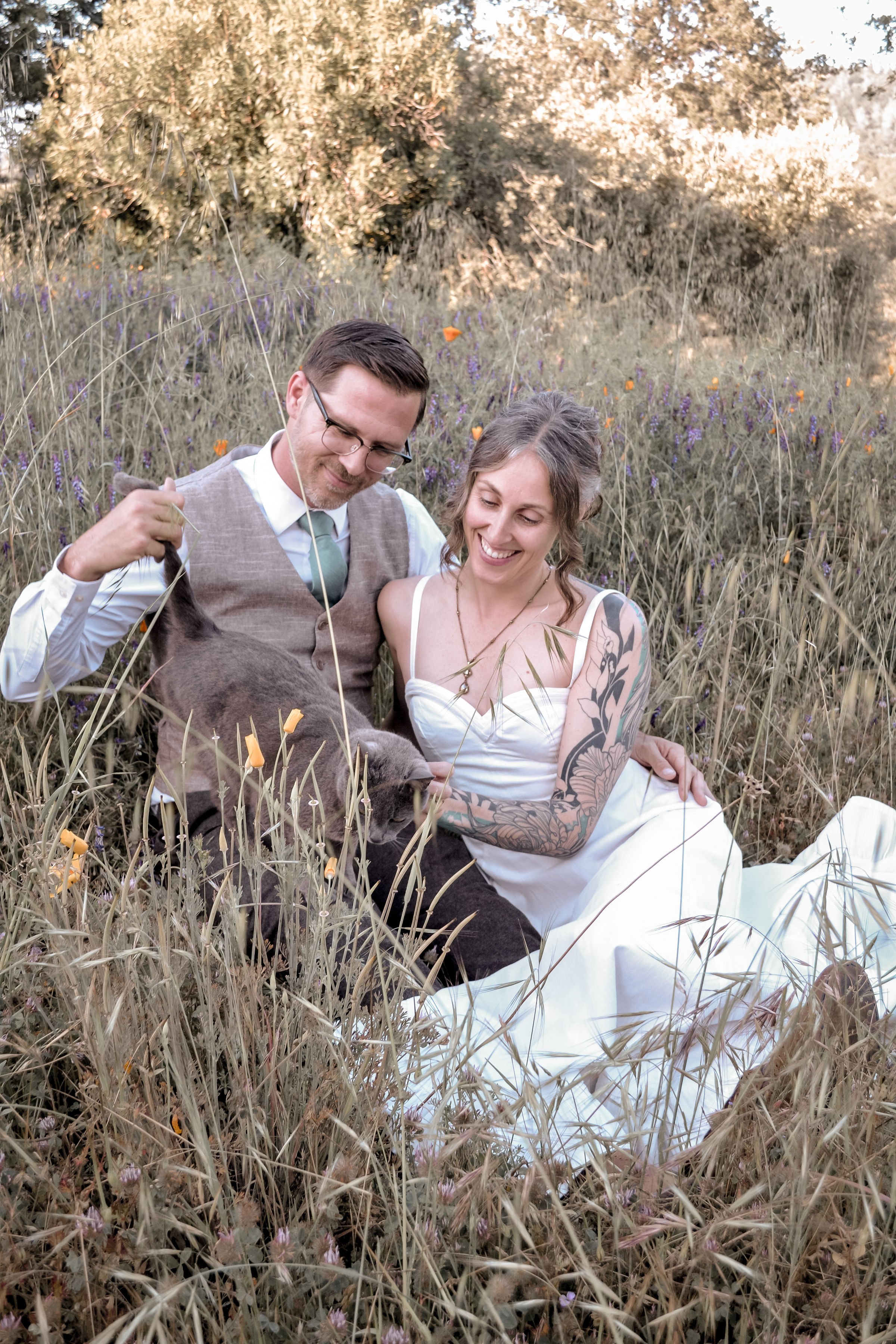 A couple sitting in a field with tall grass and wildflowers, smiling and enjoying each other's company.
