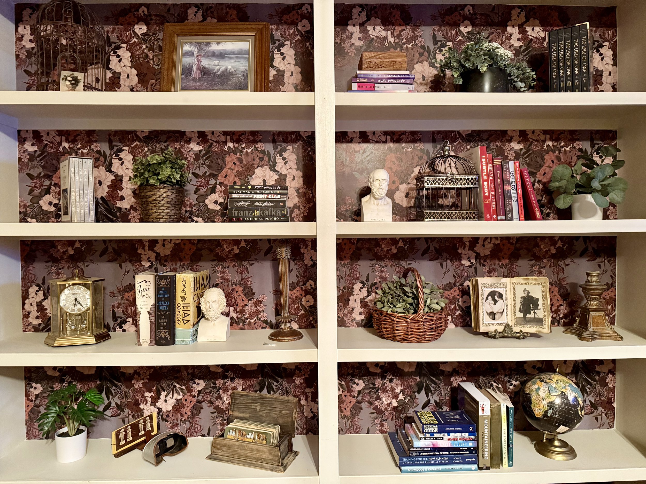 White bookshelf with five shelves, decorated with books, plants, and decorative objects against floral wallpaper background.